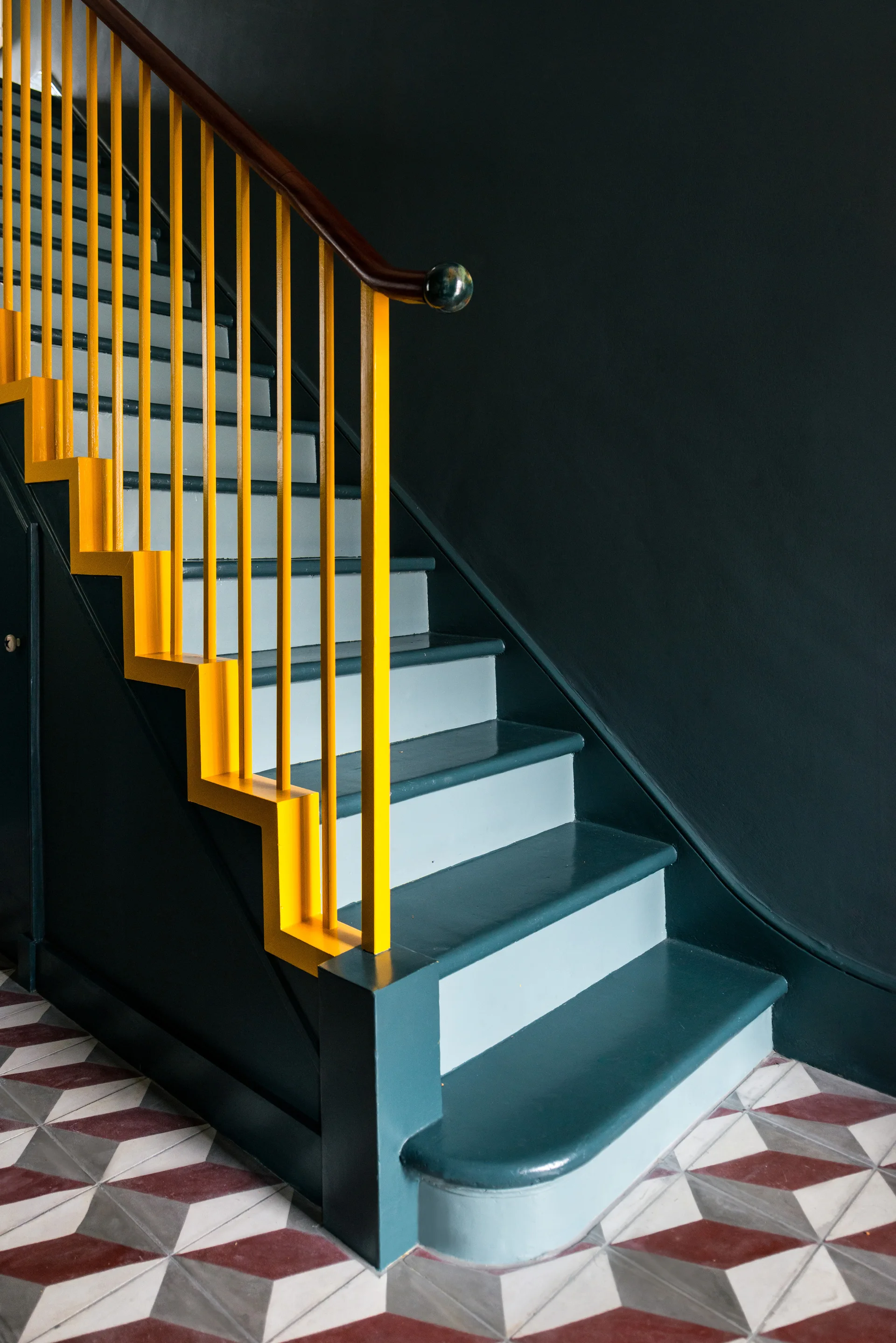 A dark teal staircase with a vibrant yellow railing climbs against a dark wall above geometric red and grey tiled flooring.