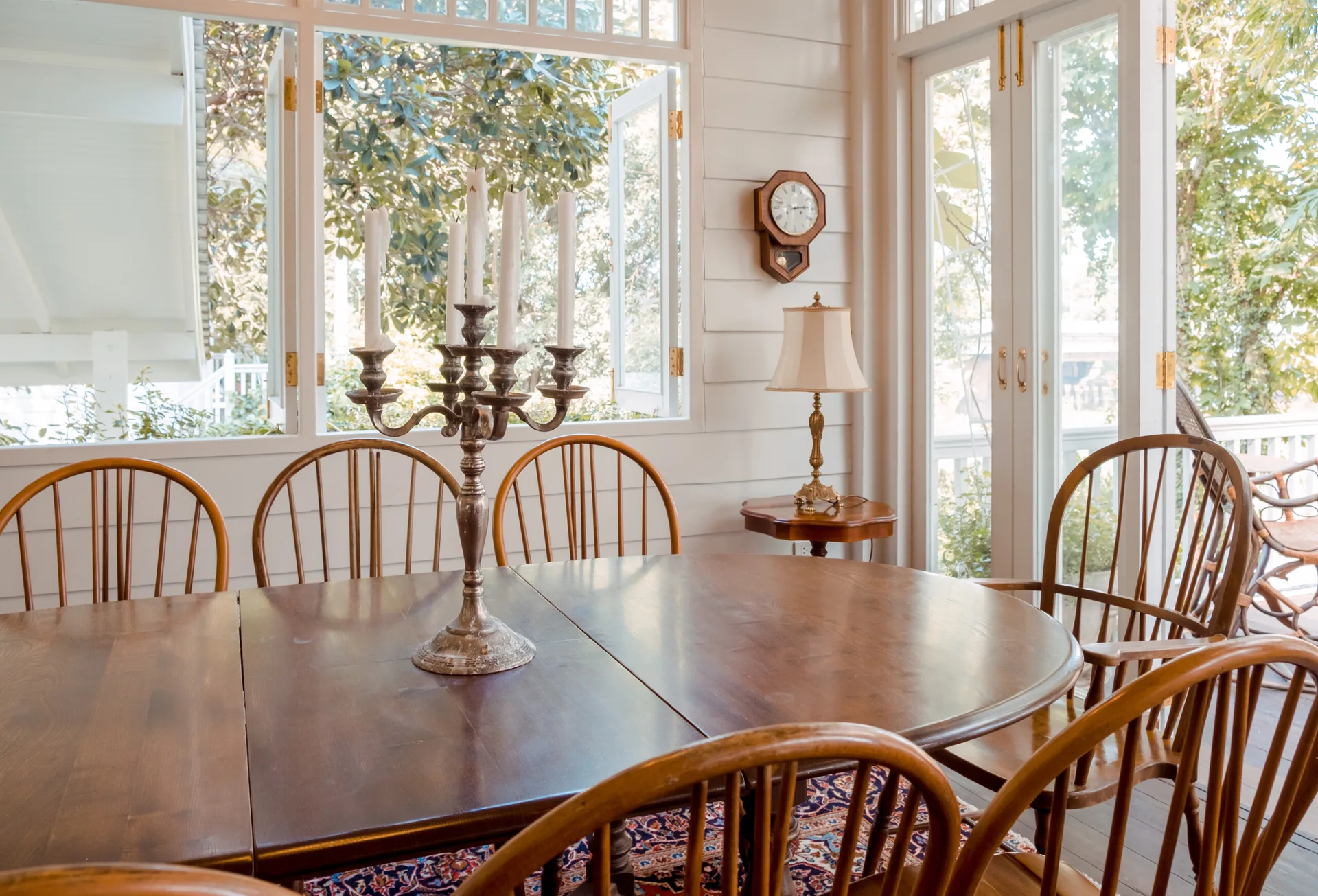 A wooden dining table with a silver candelabra centerpiece is surrounded by spindle-back chairs in a bright room with white paneled walls.
