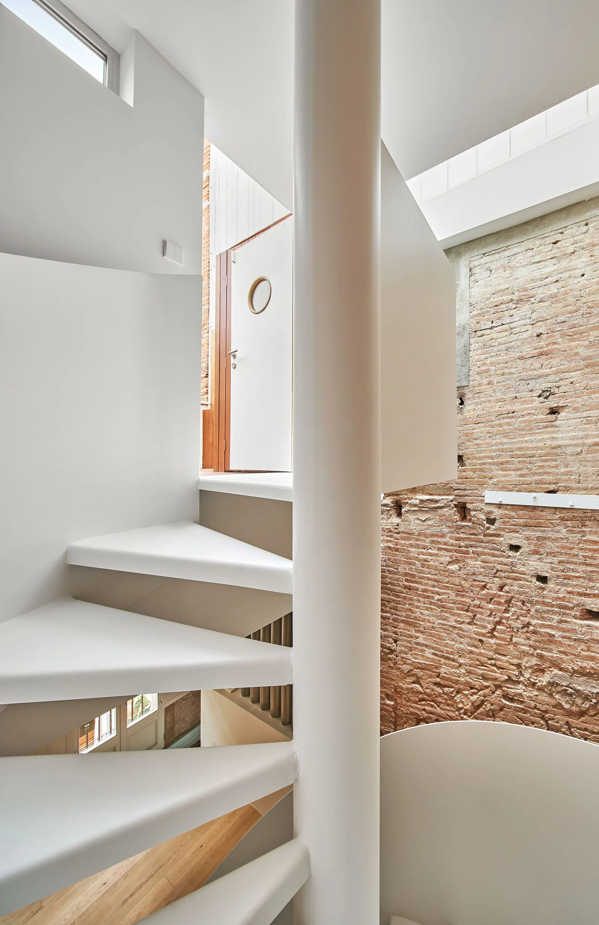 A white metal spiral staircase curves upward next to a textured red brick wall lit by a skylight.