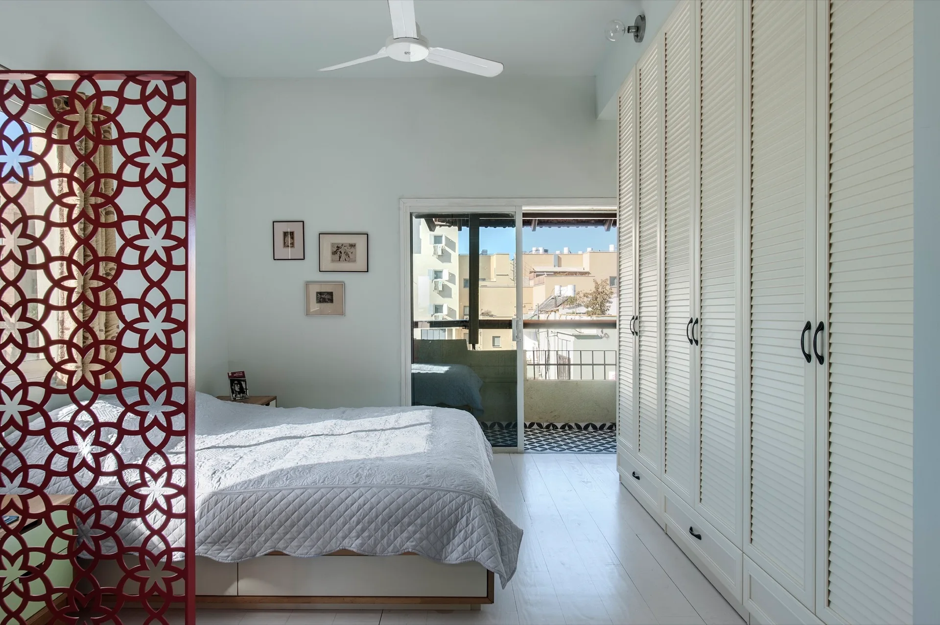 A bright white bedroom features a large louvered wardrobe cupboard, a bed with quilted bedding, and a prominent red geometric lattice screen.