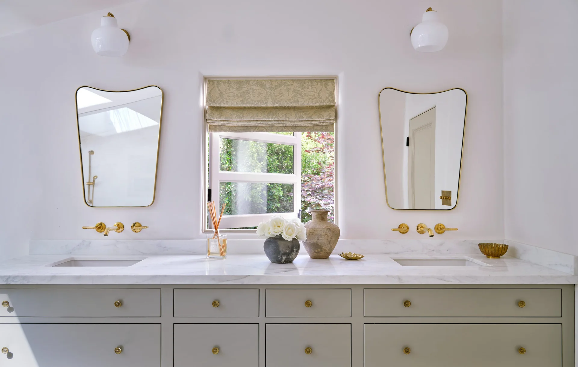 A sage green double bathroom vanity featuring a white marble countertop, wall-mounted brass faucets, curved gold-rimmed mirrors, and a central window.