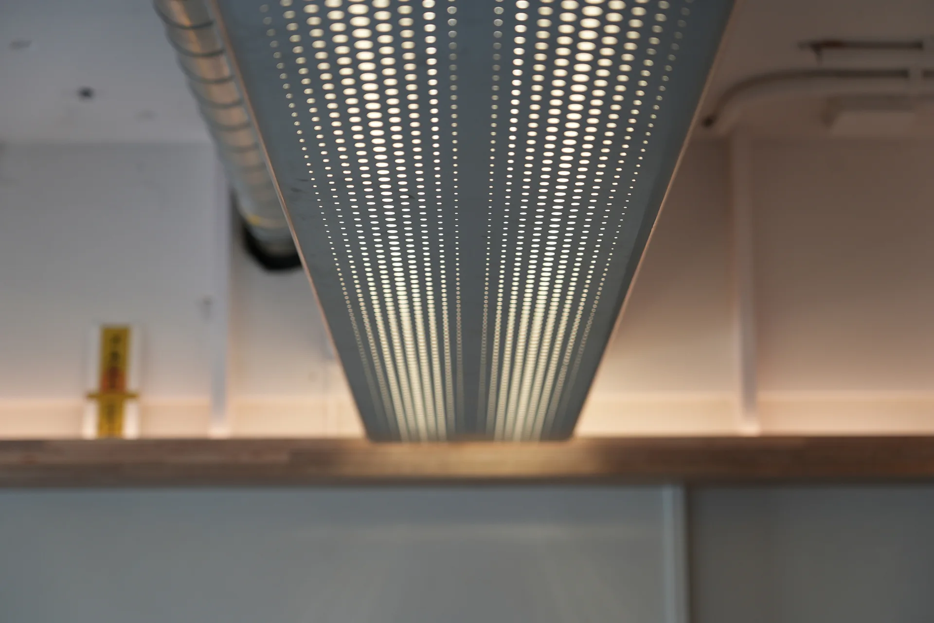 A close-up view of an elongated perforated metal pendant light fixture emitting a warm glow below a white ceiling and above a wooden shelf.