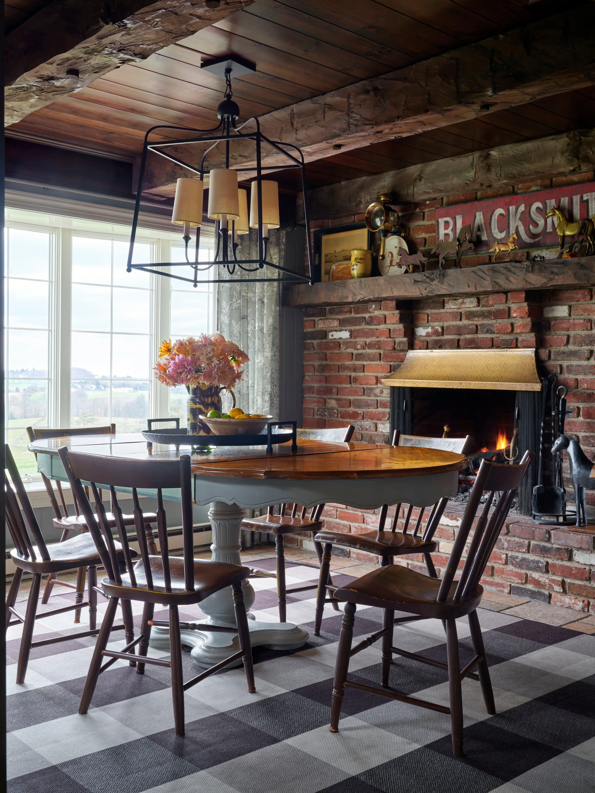 A rustic dining room showcases a wooden table and chairs on a checkered rug, situated next to a brick fireplace with a heavy wood mantel.