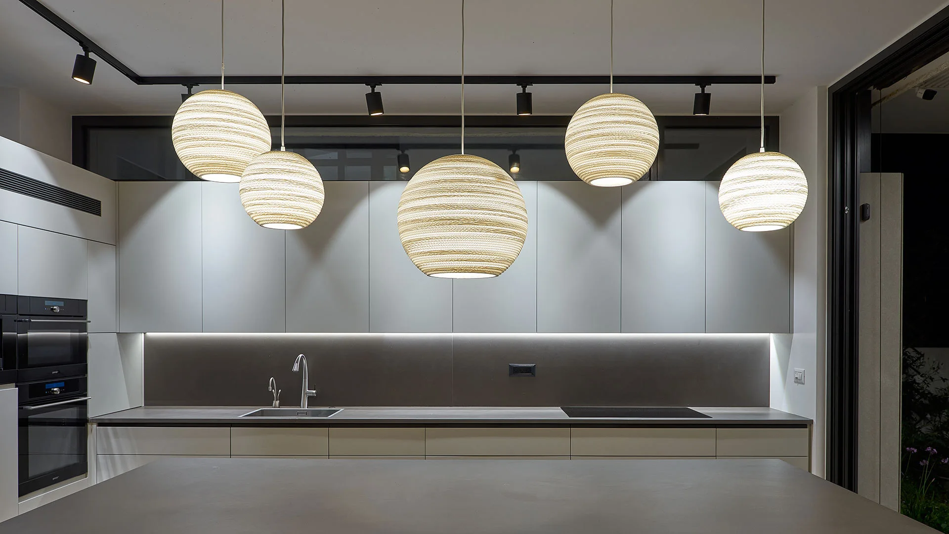 A minimalist kitchen features five glowing ribbed pendant lights hanging over a dark countertop with handleless white cabinets.