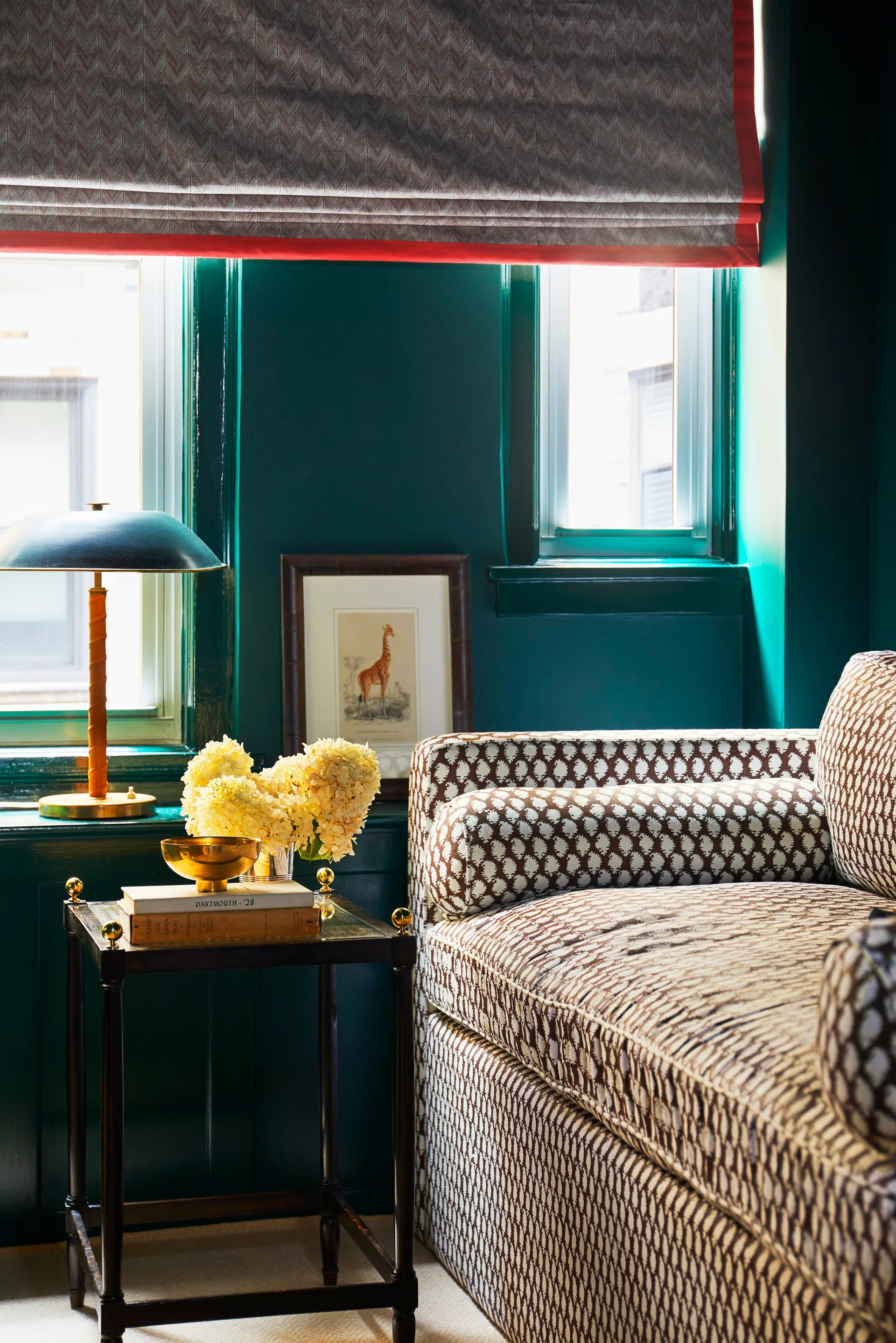 A cozy reading nook features glossy dark green walls, a patterned sofa, a wooden side table, and a herringbone Roman shade with red trim.