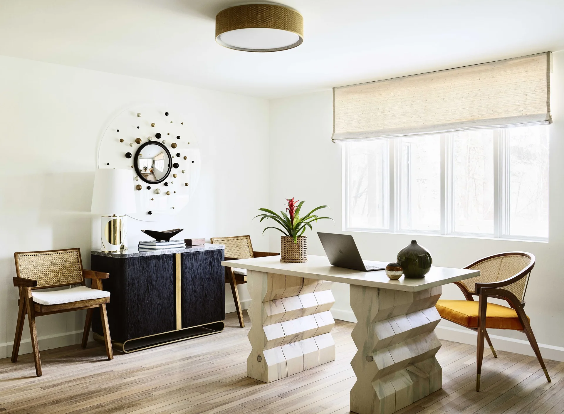 A bright home office features a sculptural wooden desk with zigzag legs, cane-back chairs, and a dark credenza under a decorative round mirror.