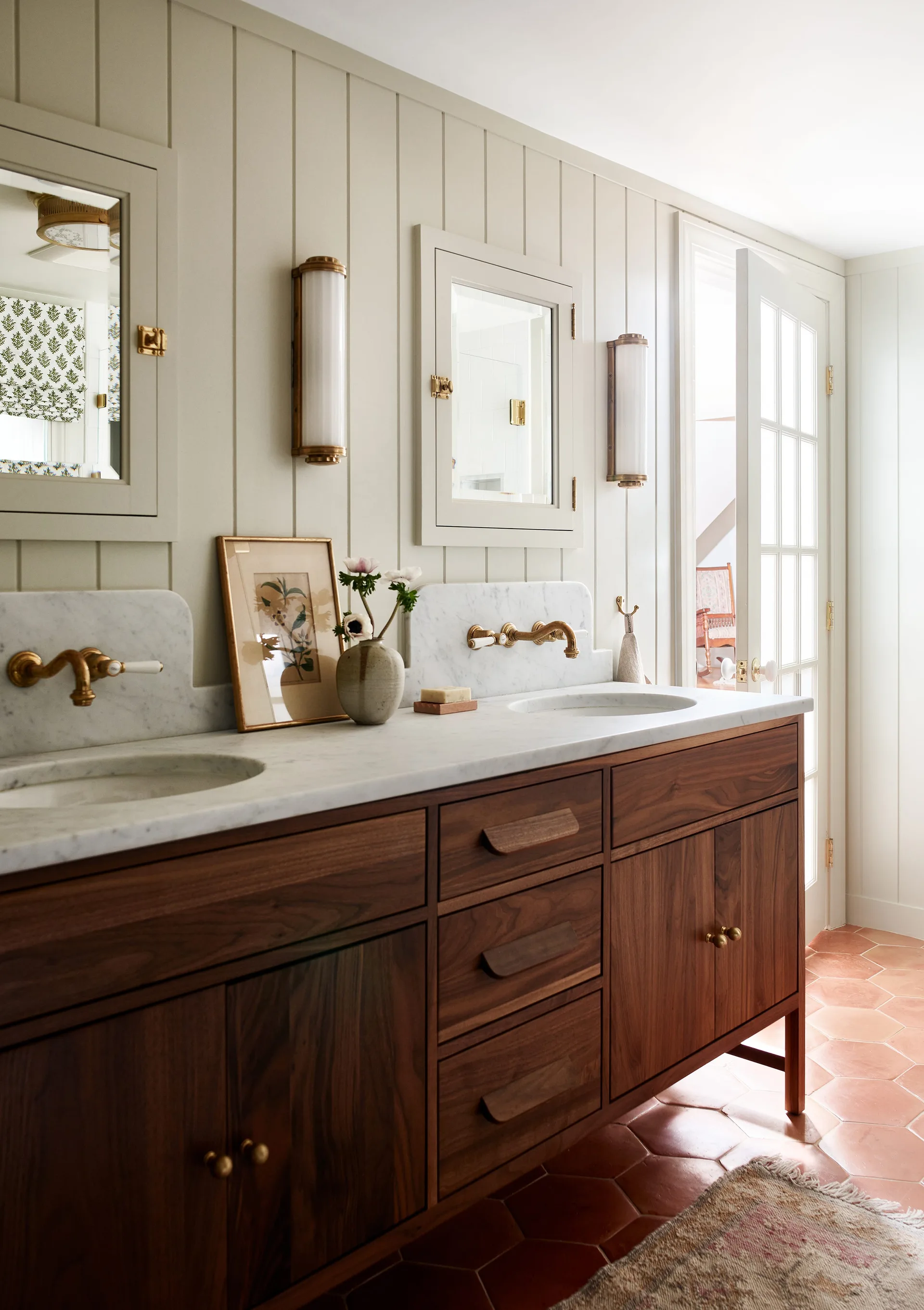 A bathroom featuring a walnut double vanity with marble countertops, wall-mounted brass faucets, vertical wood paneling, and hexagonal terracotta floor tiles.