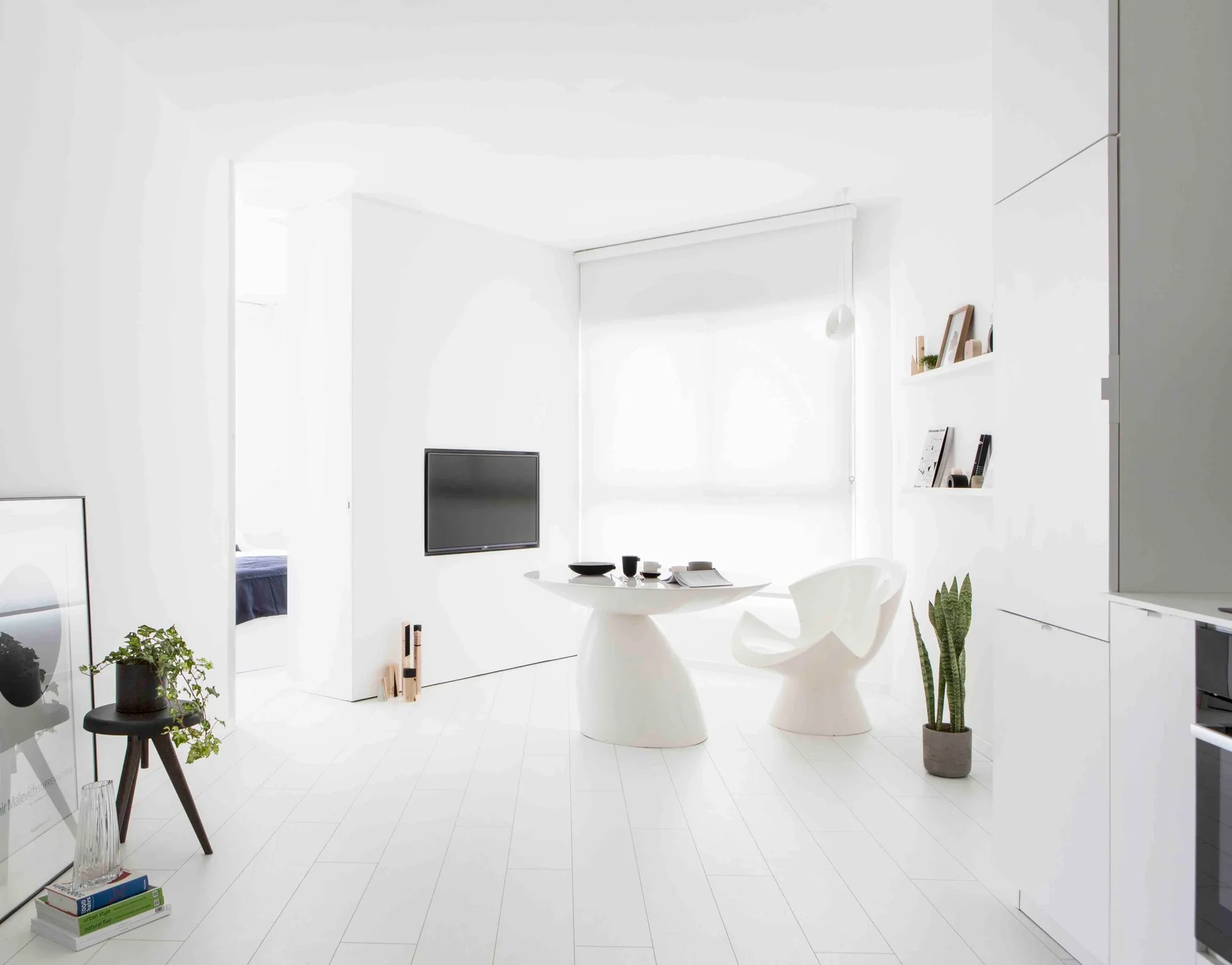A bright white minimalist living area features a curved sculptural pedestal table, a molded plastic armchair, and white wood plank flooring.