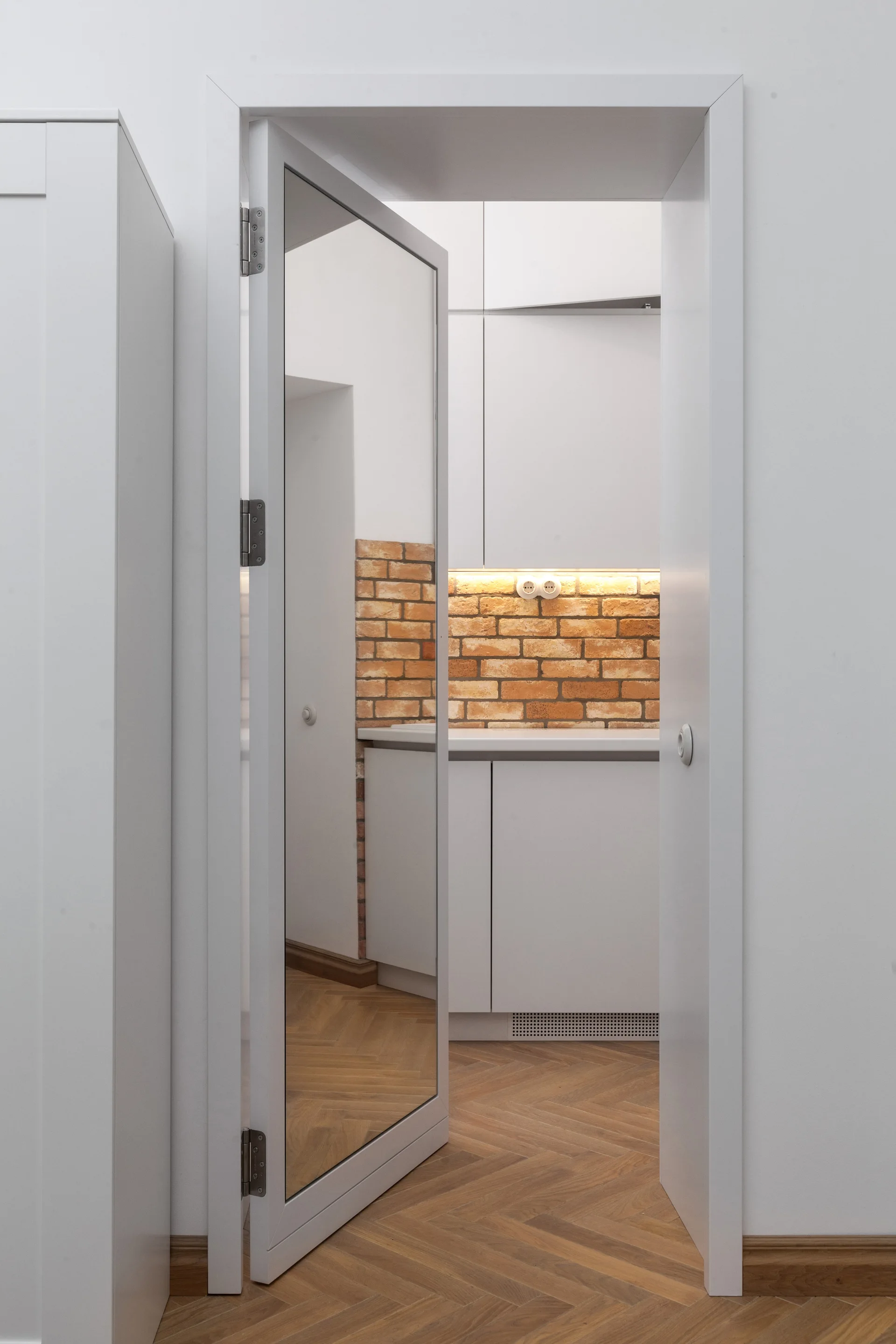 An open white door with a full-length mirror panel looks into a kitchen with white cabinets, a brick backsplash, and herringbone floors.