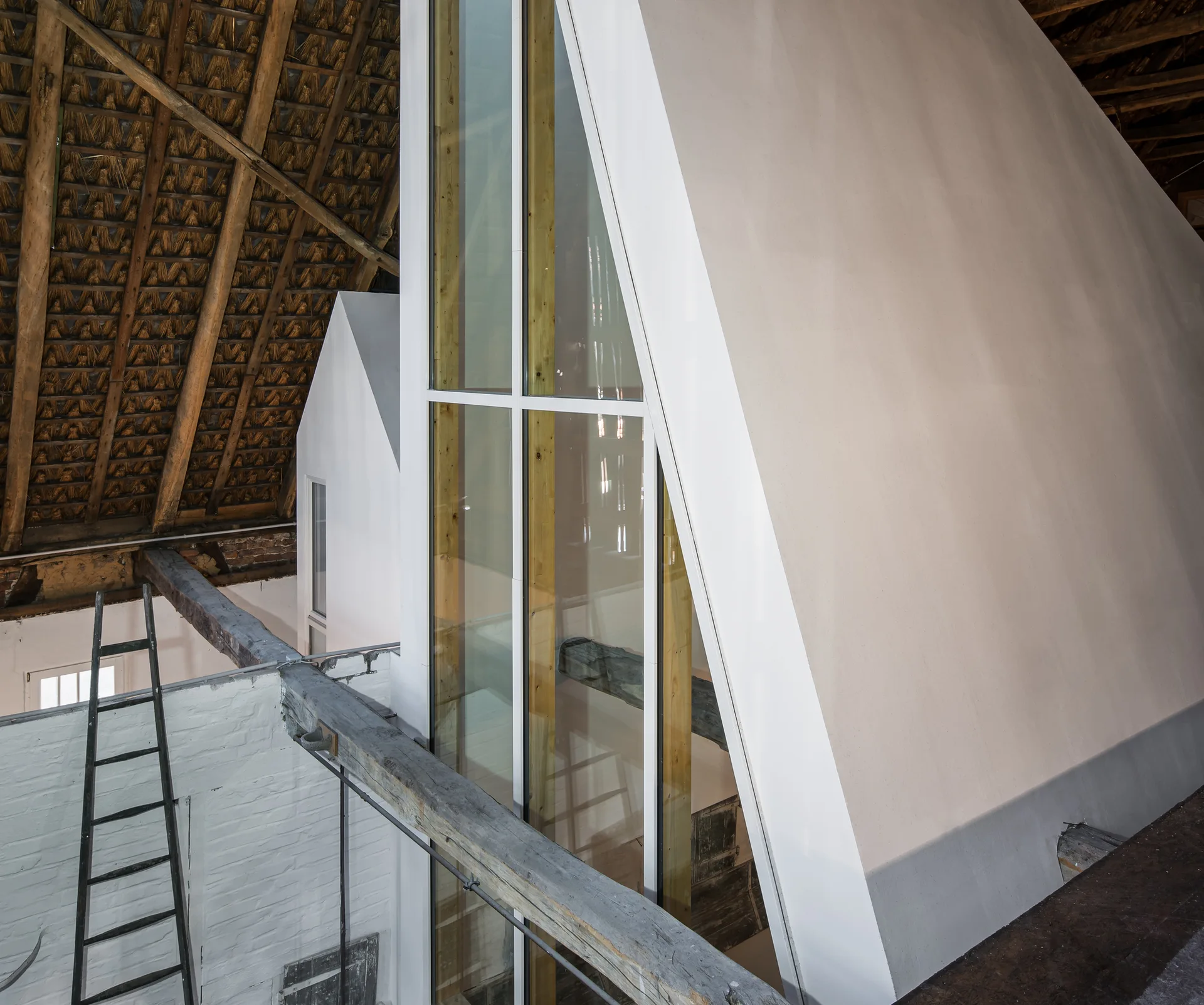 An attic space featuring an exposed thatch ceiling, rustic wood beams, a black ladder, and a contemporary white triangular structure with tall glass windows.