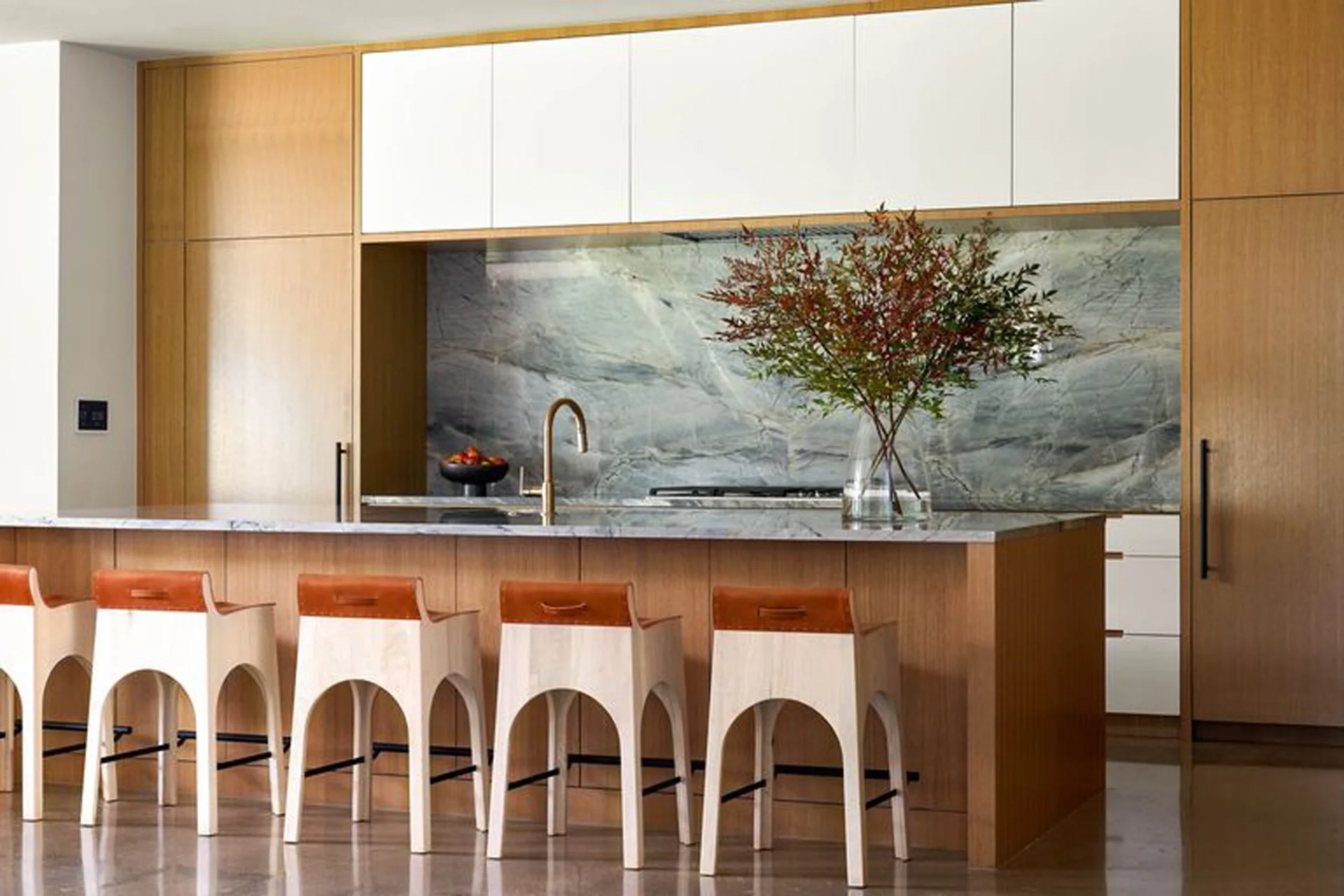 A modern kitchen features a wood island with four arched wooden bar stools, white upper cabinets, and a gray veined stone slab backsplash.