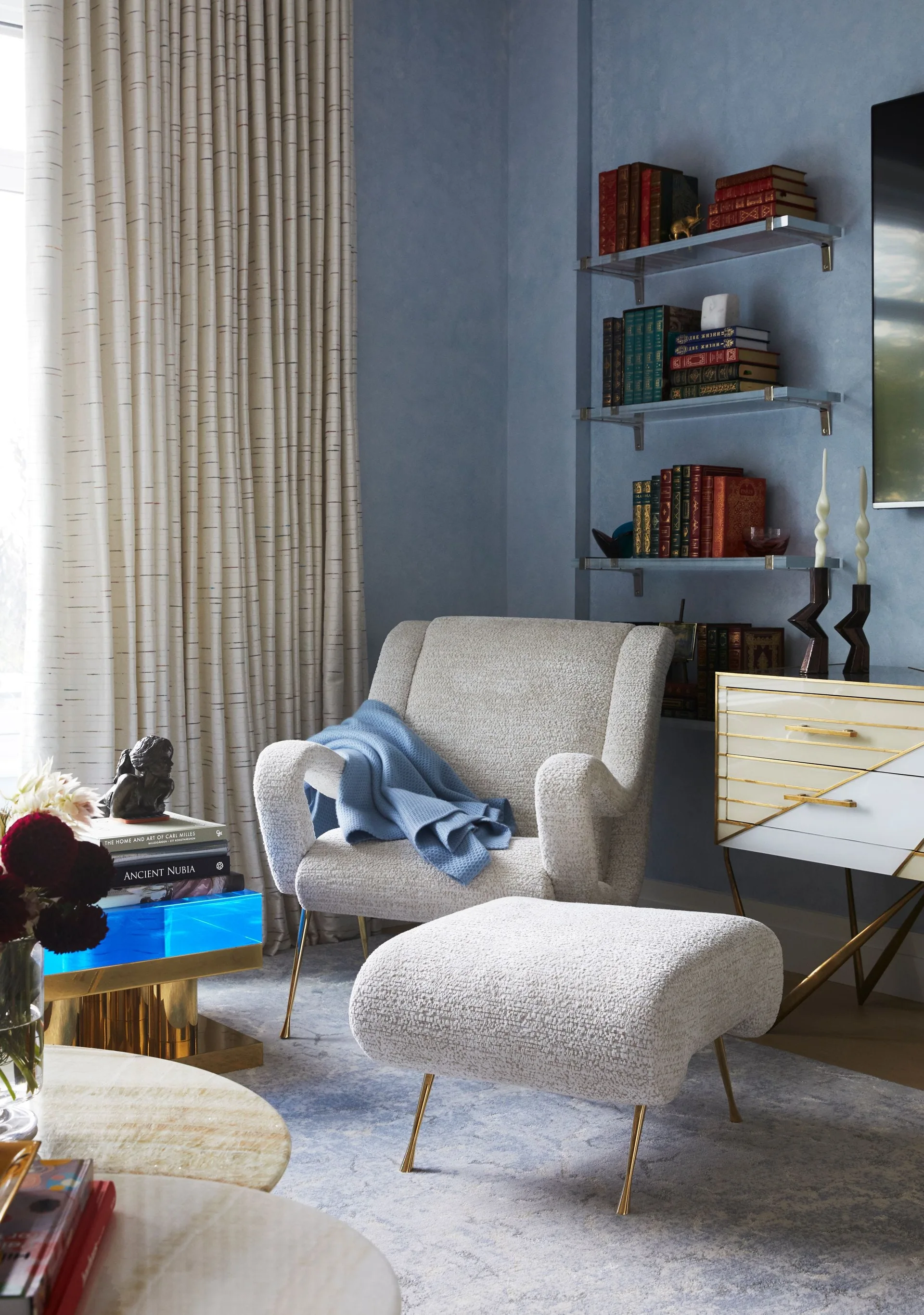 A textured cream armchair and matching ottoman rest in a blue room next to pleated curtains and recessed glass shelving.