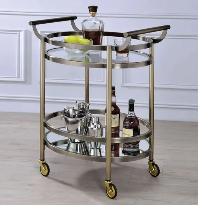 Two-tiered oval bar cart in brushed gold with glass shelves holding liquor bottles and bar tools against a white paneled wall.