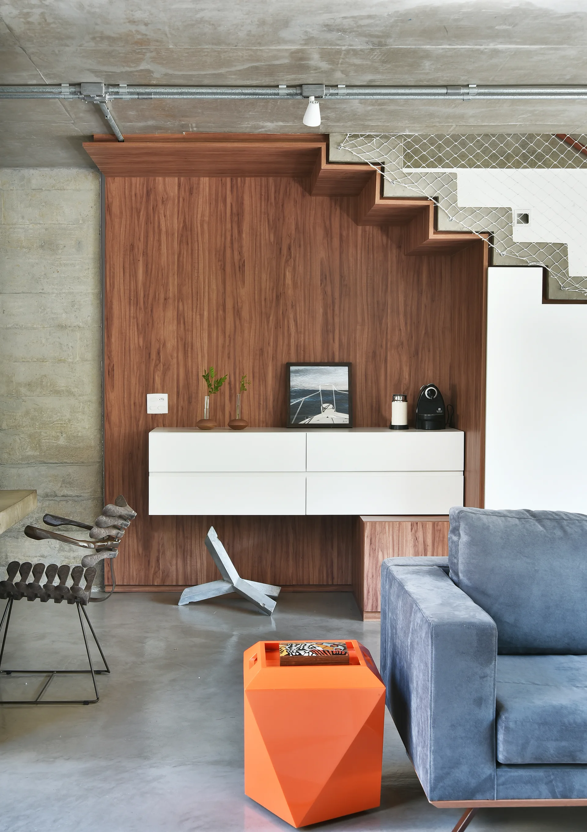 A blue sofa and orange geometric stool stand near a floating white cabinet mounted on a wood-paneled wall under a staircase with safety netting.