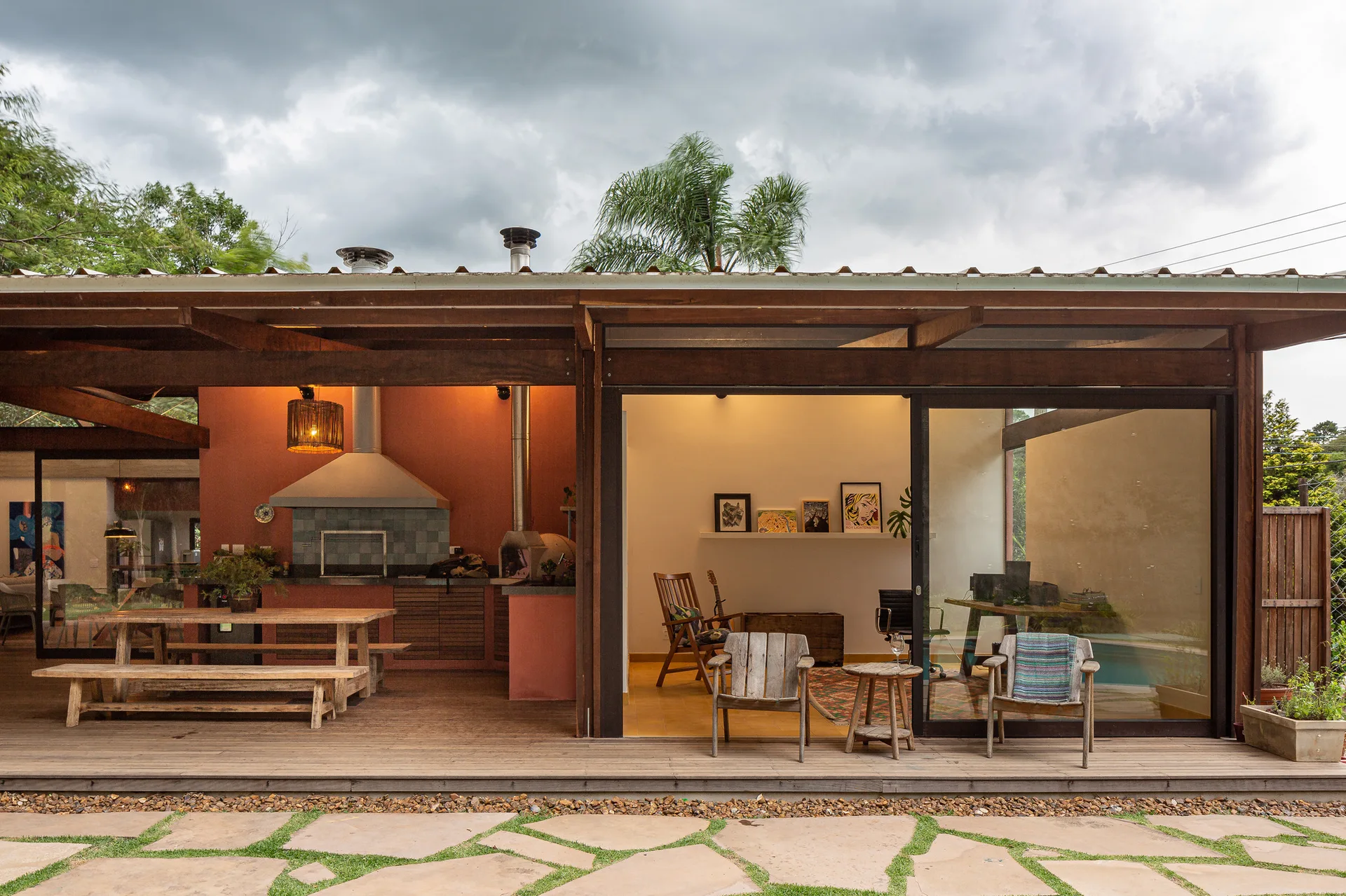 A covered wooden deck patio featuring a built-in kitchen against a terracotta wall, a rustic dining table, and glass sliding doors leading indoors.