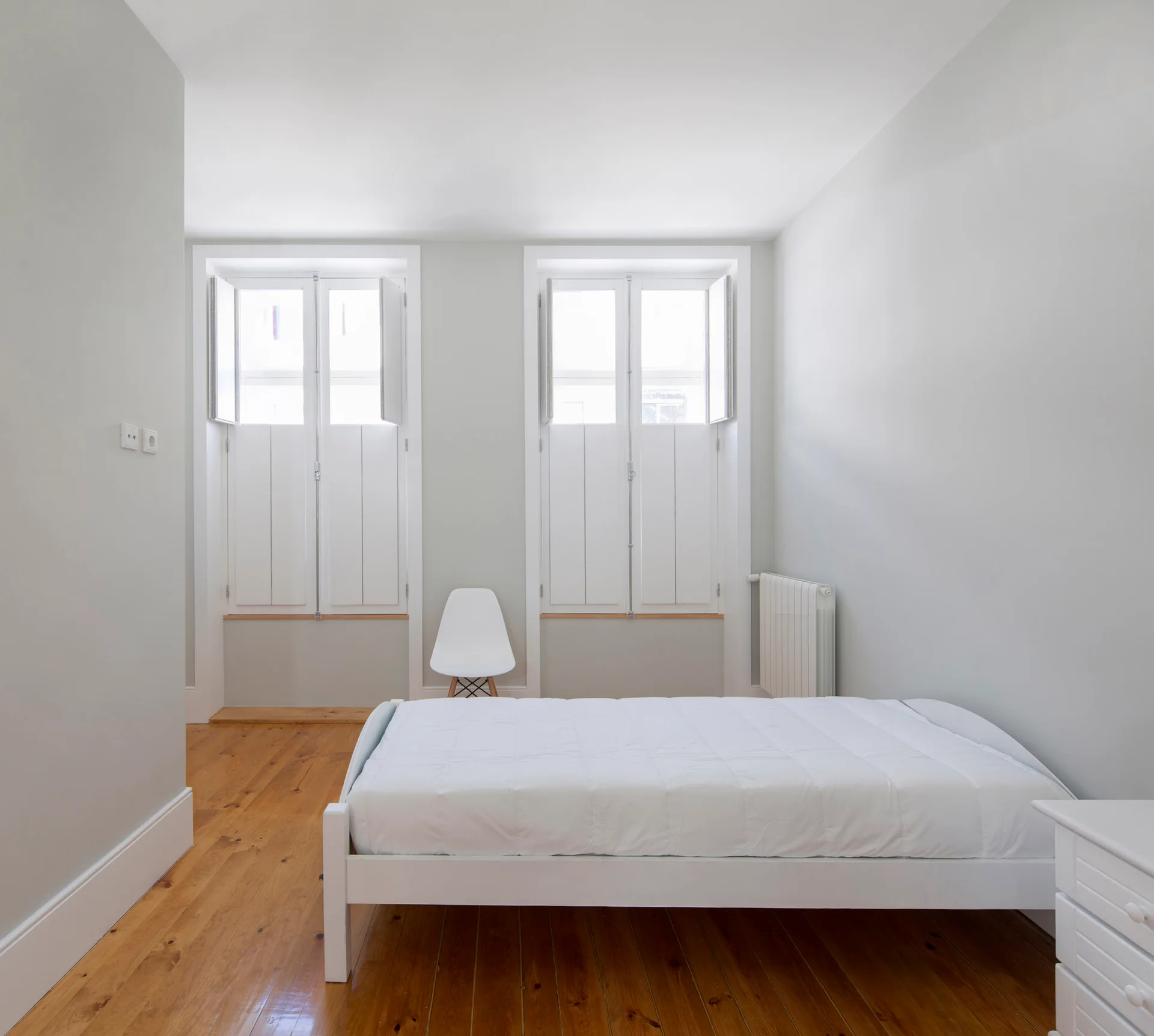 A minimalist bedroom features a low white bed on pine wood floors beneath two tall windows with integrated white shutters.