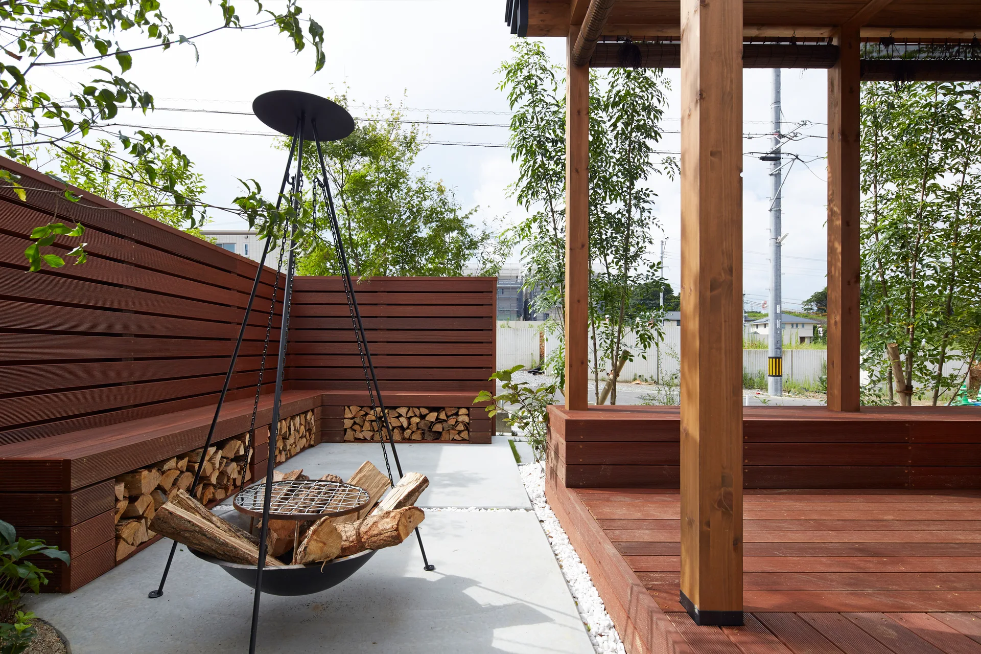 A modern outdoor patio features a black tripod fire pit on a concrete floor next to a wooden deck, a privacy fence, and a bench with integrated log storage.