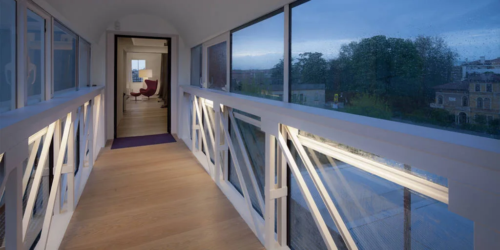 A minimalist glass corridor features light wood flooring, an arched ceiling, and white geometric railings illuminated by integrated linear lights.