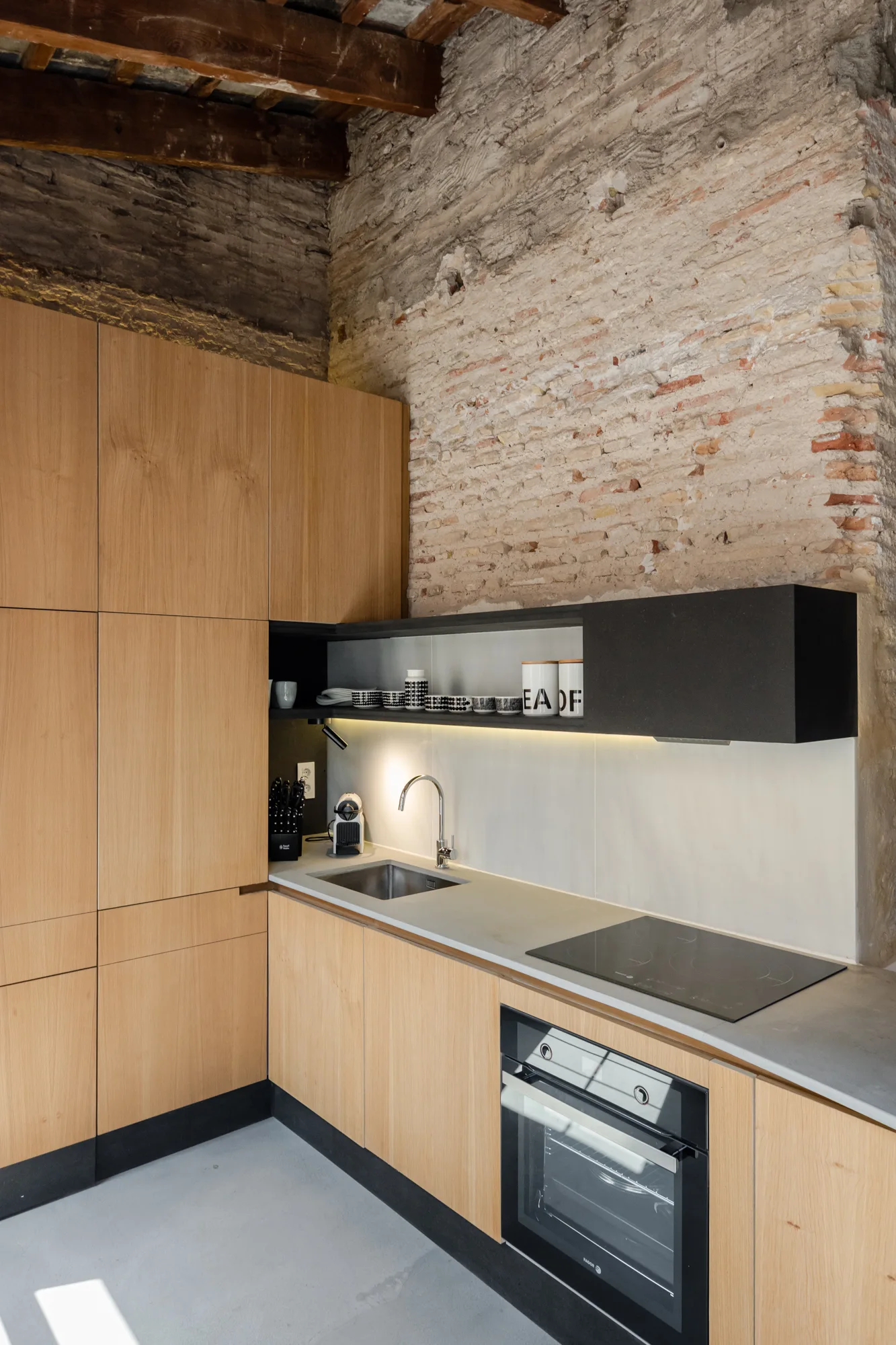 A modern kitchen with wood cabinets, concrete countertops, a black floating shelf, and a textured exposed brick wall.