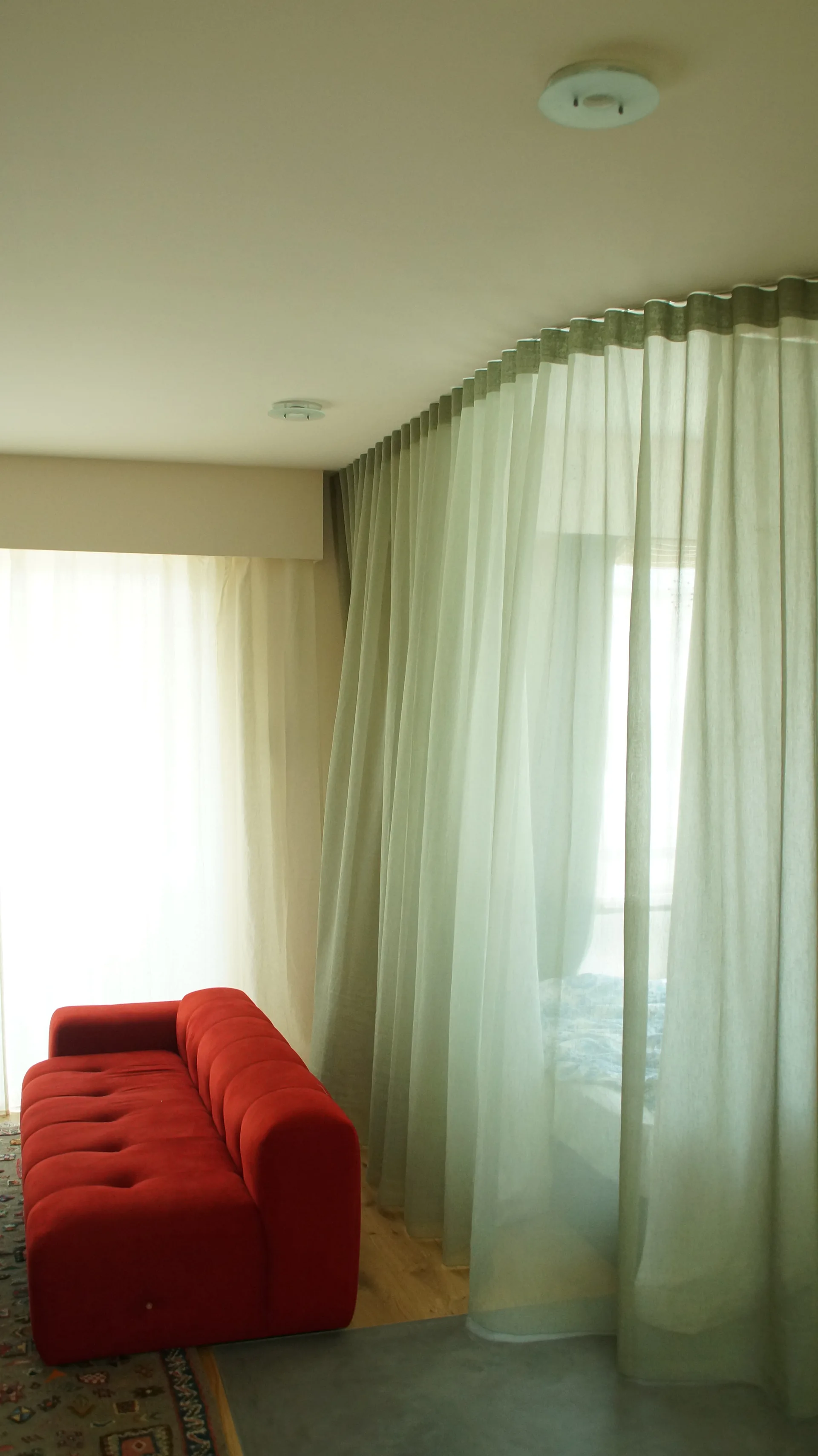 A vibrant red tufted sofa sits next to floor-to-ceiling sheer sage green curtains suspended from a curved ceiling track.