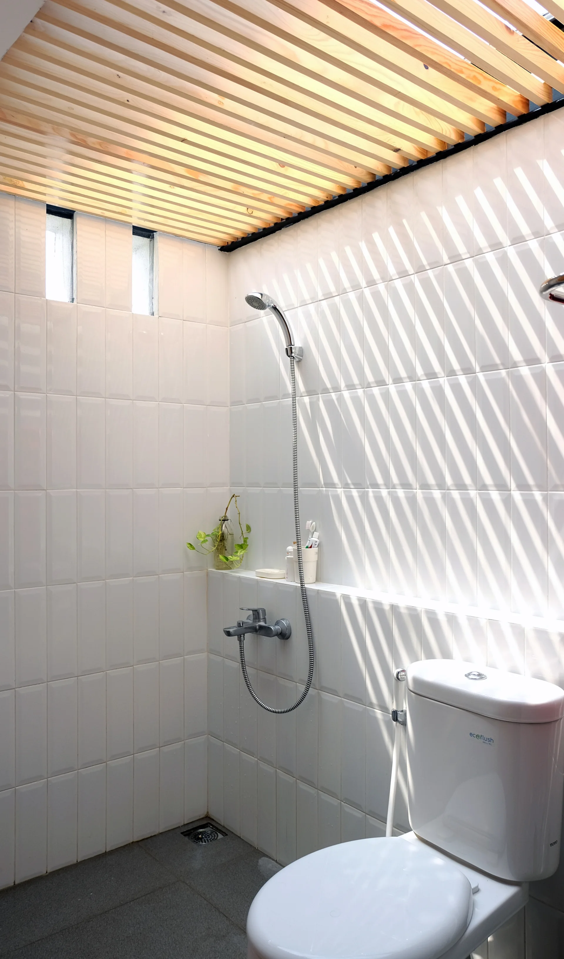 A minimalist bathroom featuring white tiled walls, a natural wood slat ceiling, a chrome shower fixture, and a white toilet.