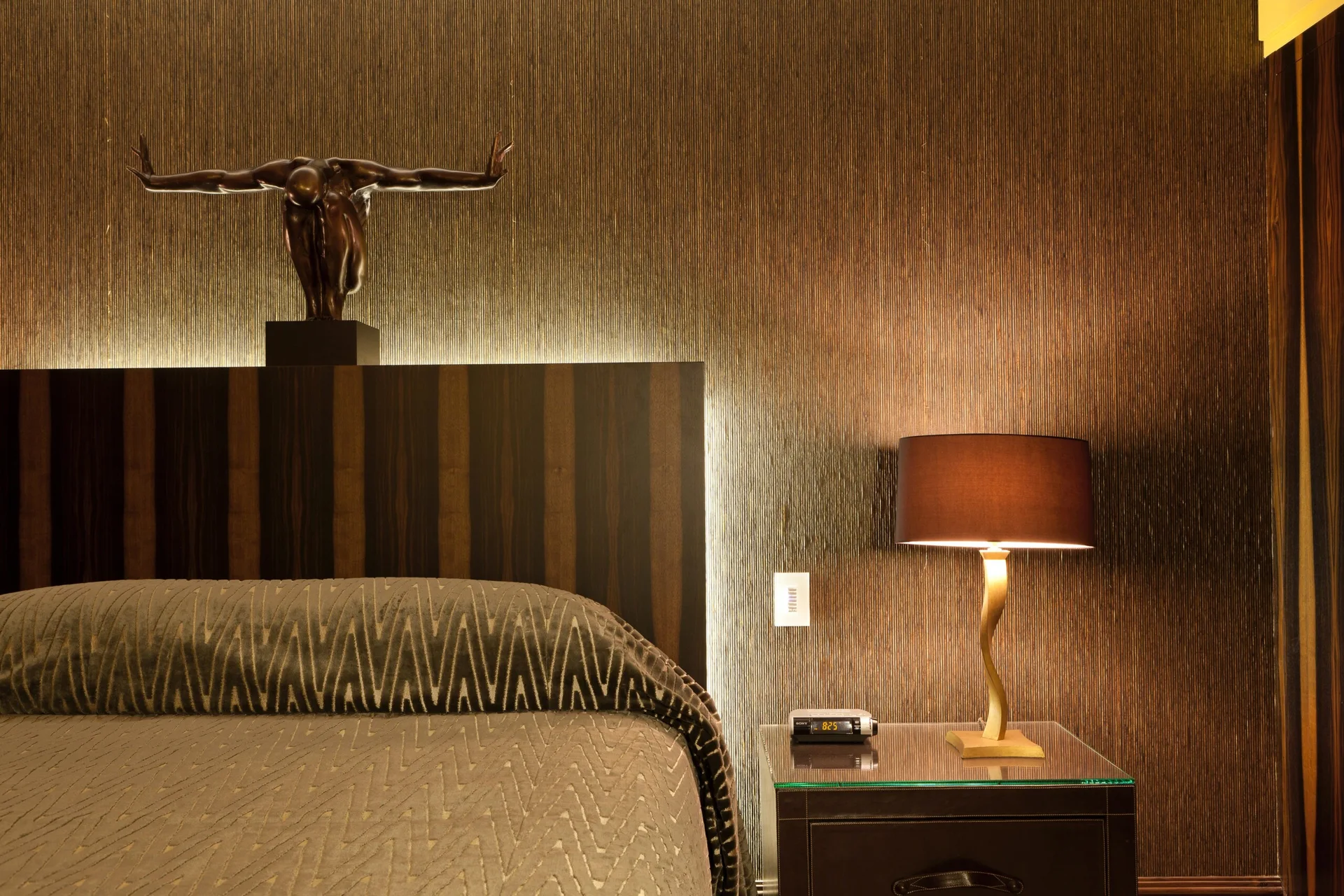 A dimly lit bedroom corner features a backlit wooden headboard beneath a bronze sculpture, a textured brown wall, and a gold lamp on a dark nightstand.