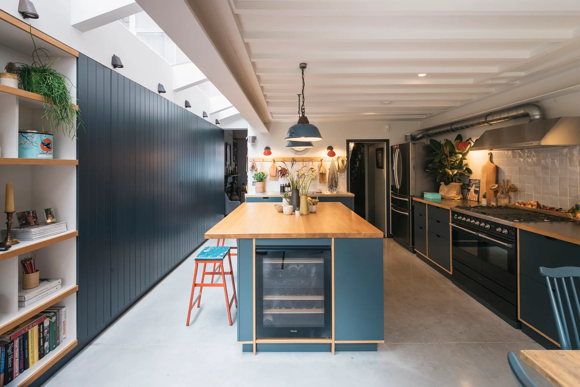 A modern kitchen features a dark blue island with a wood countertop, integrated wine fridge, blue cabinetry, concrete floor, and skylight.