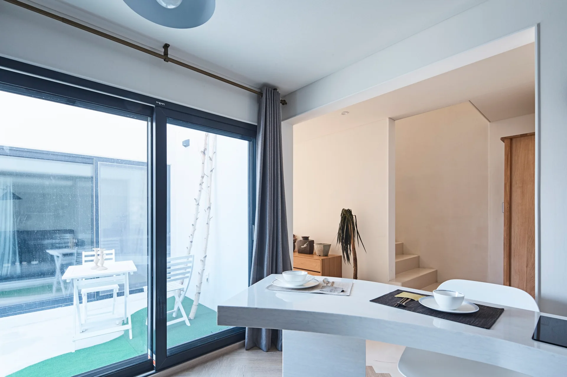 A minimalist dining room features a glossy white table set with bowls, looking out through large sliding glass doors to a small white patio.