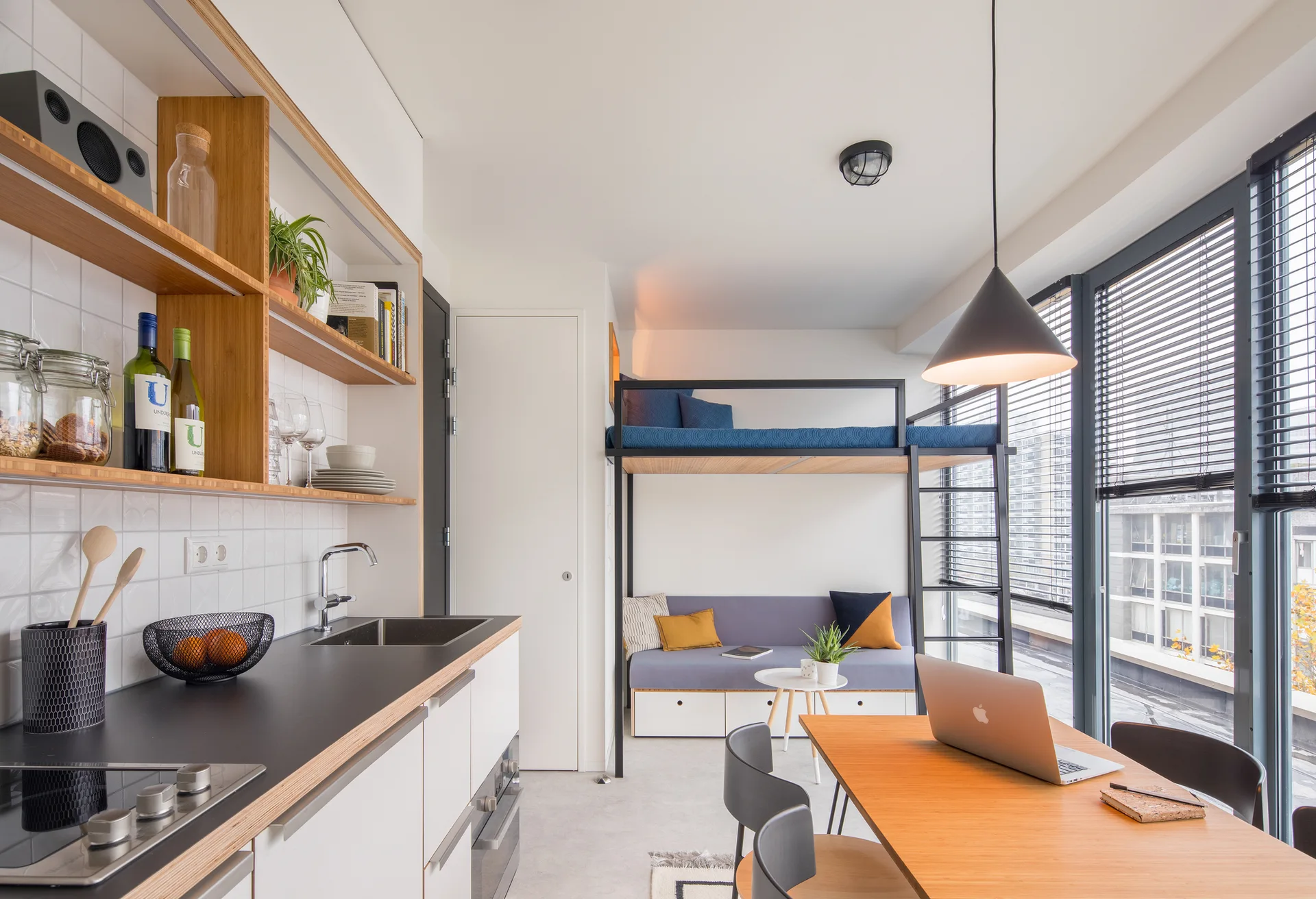 A modern studio apartment featuring a kitchen counter with wooden shelves, a metal loft bed over a gray sofa, and a dining table by large windows.