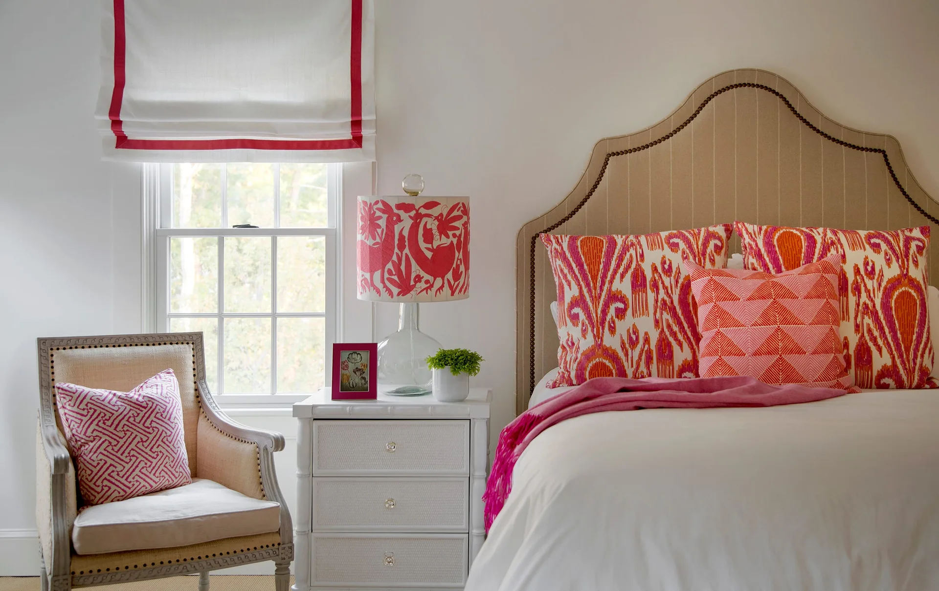 A bedroom featuring a beige upholstered headboard, white nightstand, patterned pink pillows, and a matching patterned table lamp next to a window.