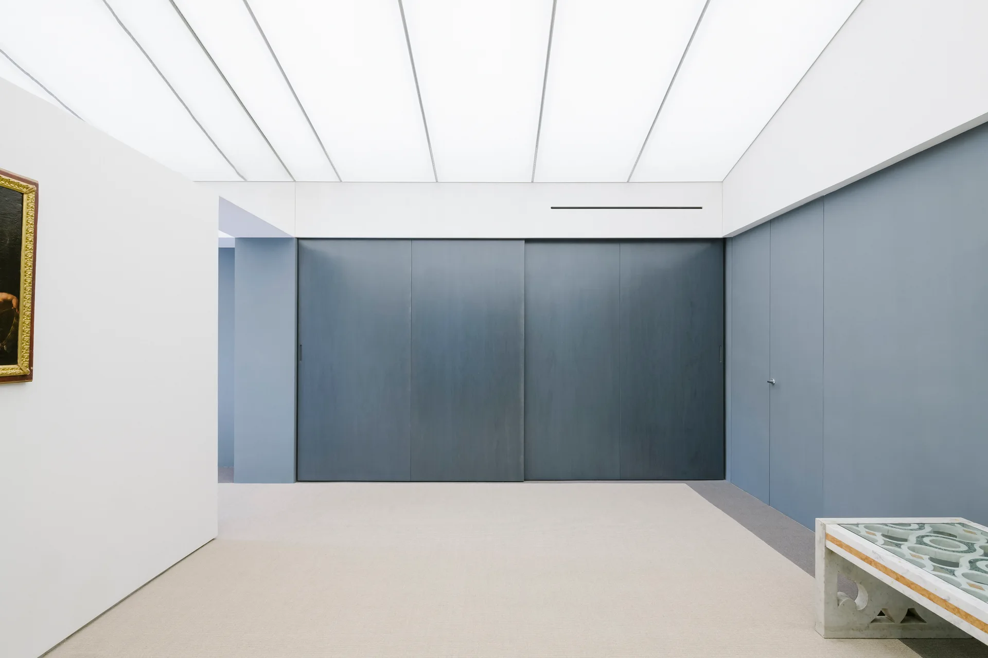 A minimalist hallway features slate blue paneled walls, a large sliding door, a low stone console, and a bright translucent grid ceiling.