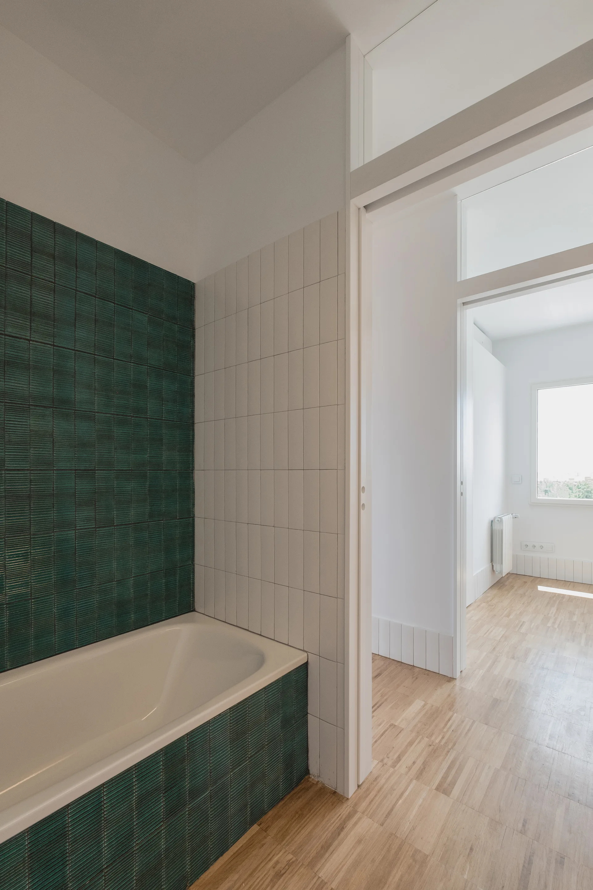 A modern bathroom features a bathtub clad in green textured tiles next to vertical white tiles with a doorway leading to a sunlit hallway.