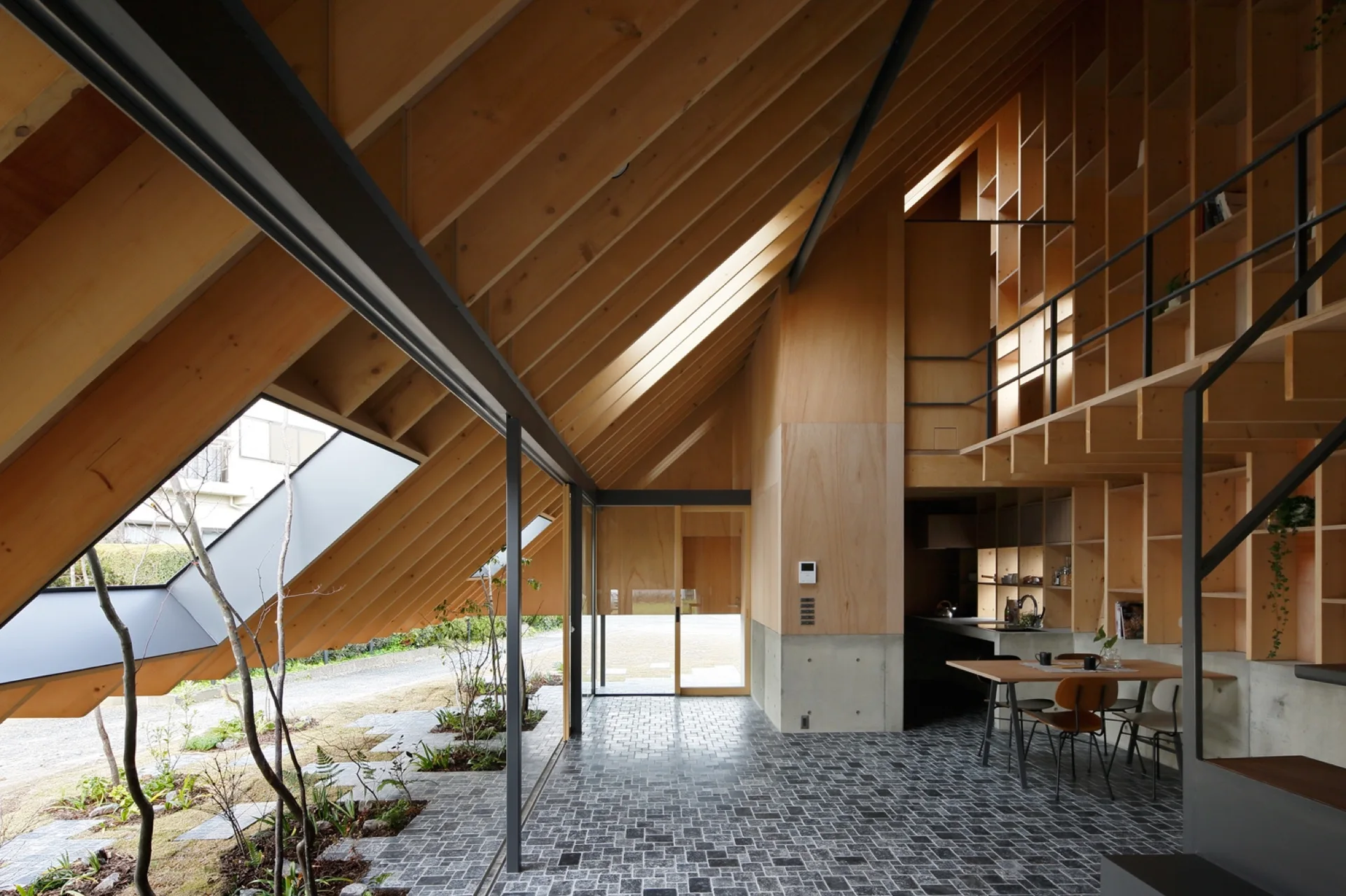 A modern residential interior with a steep pitched wood ceiling, exposed rafters, built-in shelving, concrete wall bases, and a dark stone tile floor.