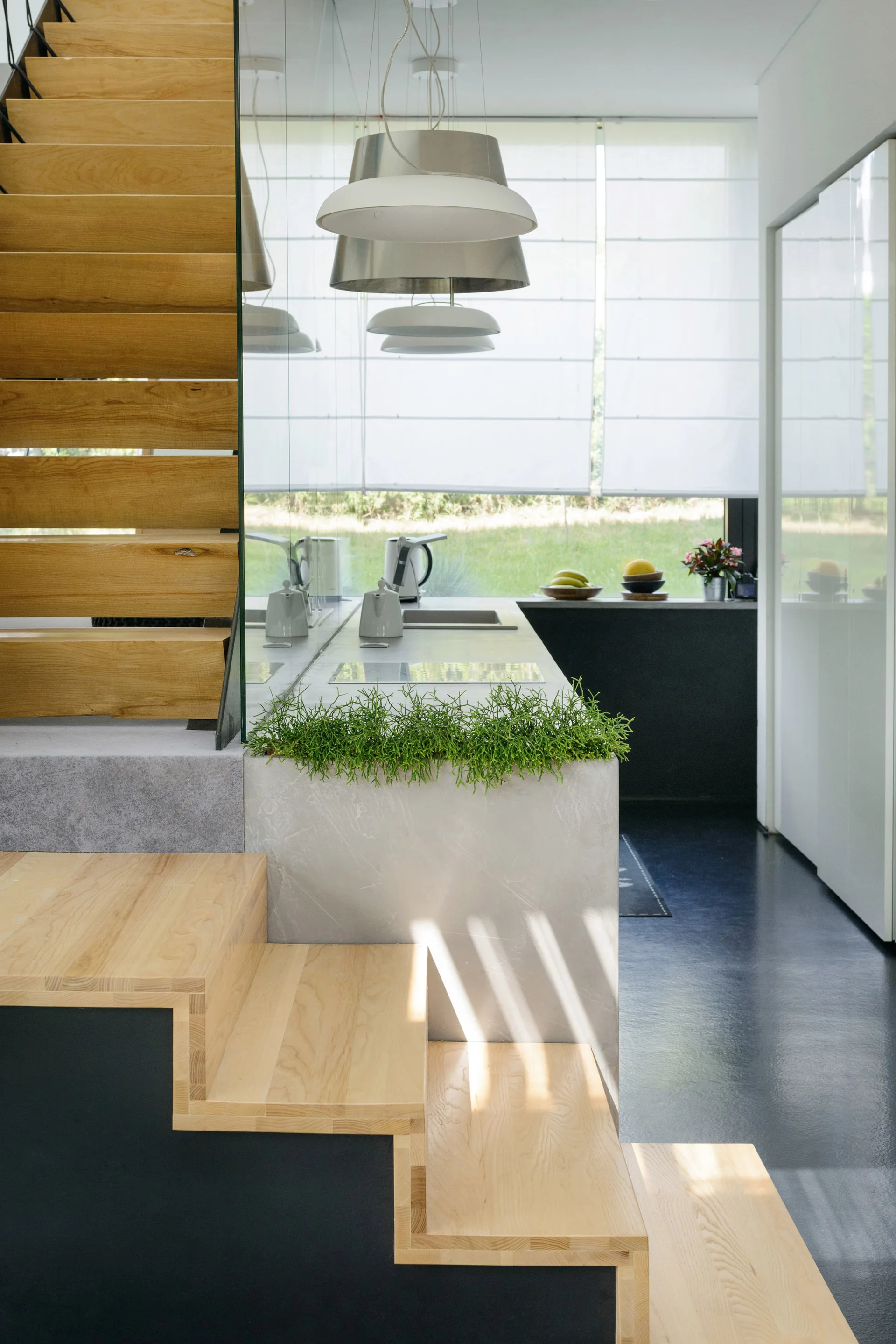 A modern kitchen interior featuring a concrete island next to a light wood open staircase, under natural light from a large window.