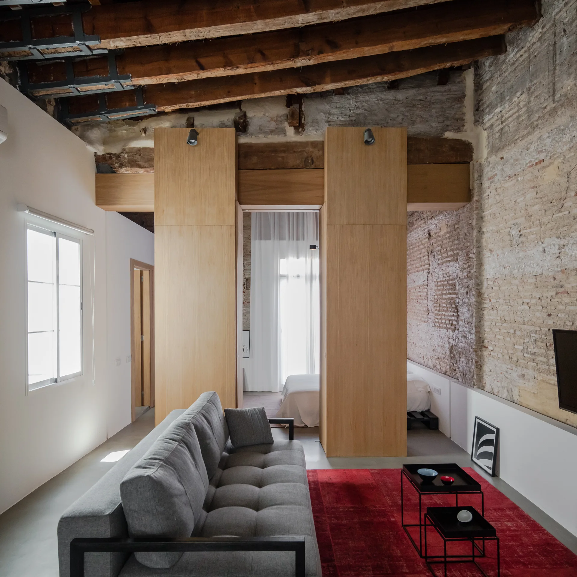 A grey tufted sofa sits on a red rug in an industrial living room featuring exposed brick walls, timber ceiling beams, and tall wood partitions.