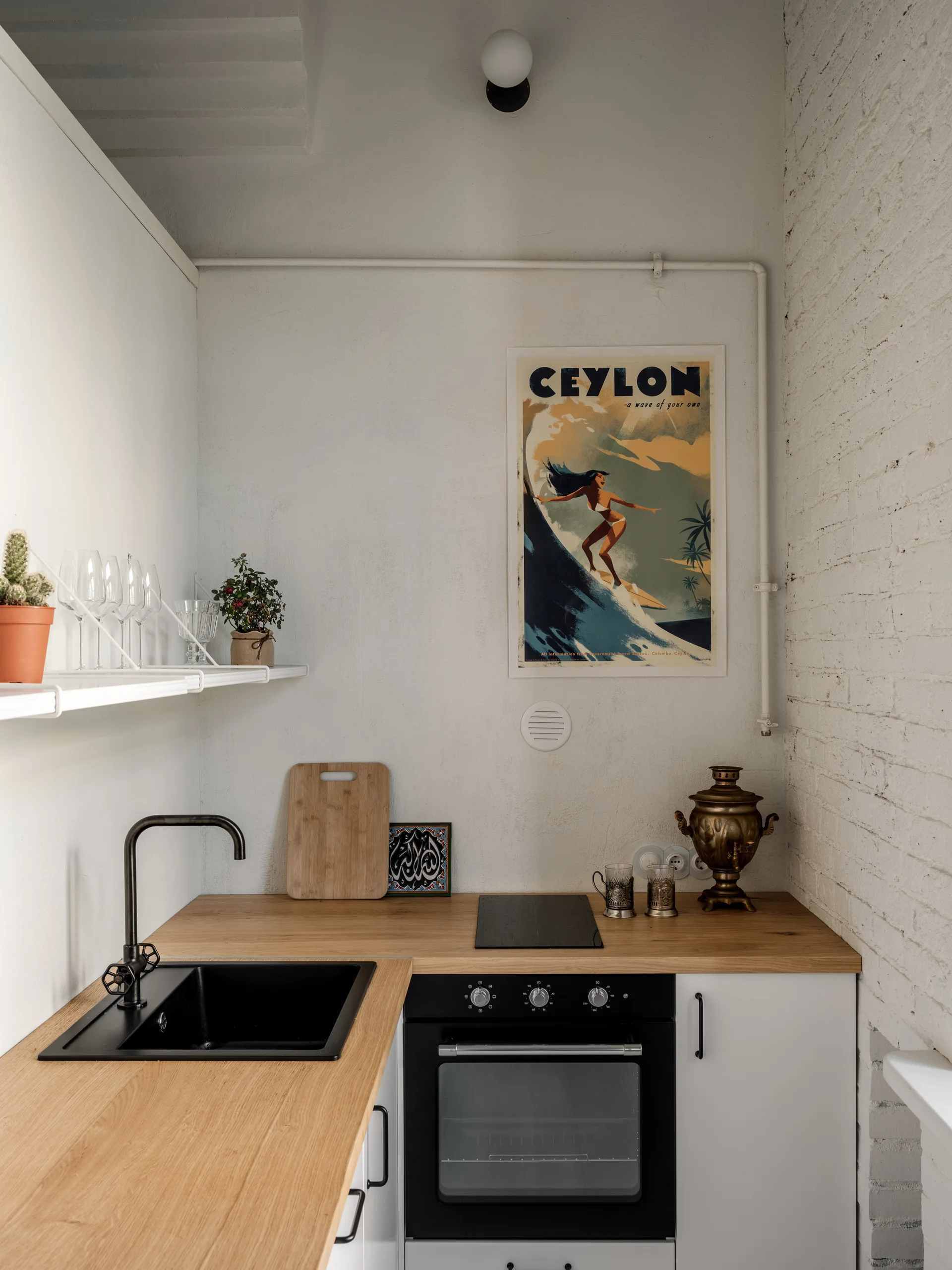 A compact white kitchenette features a light wood countertop, black sink, integrated oven, and a framed travel poster on the wall.