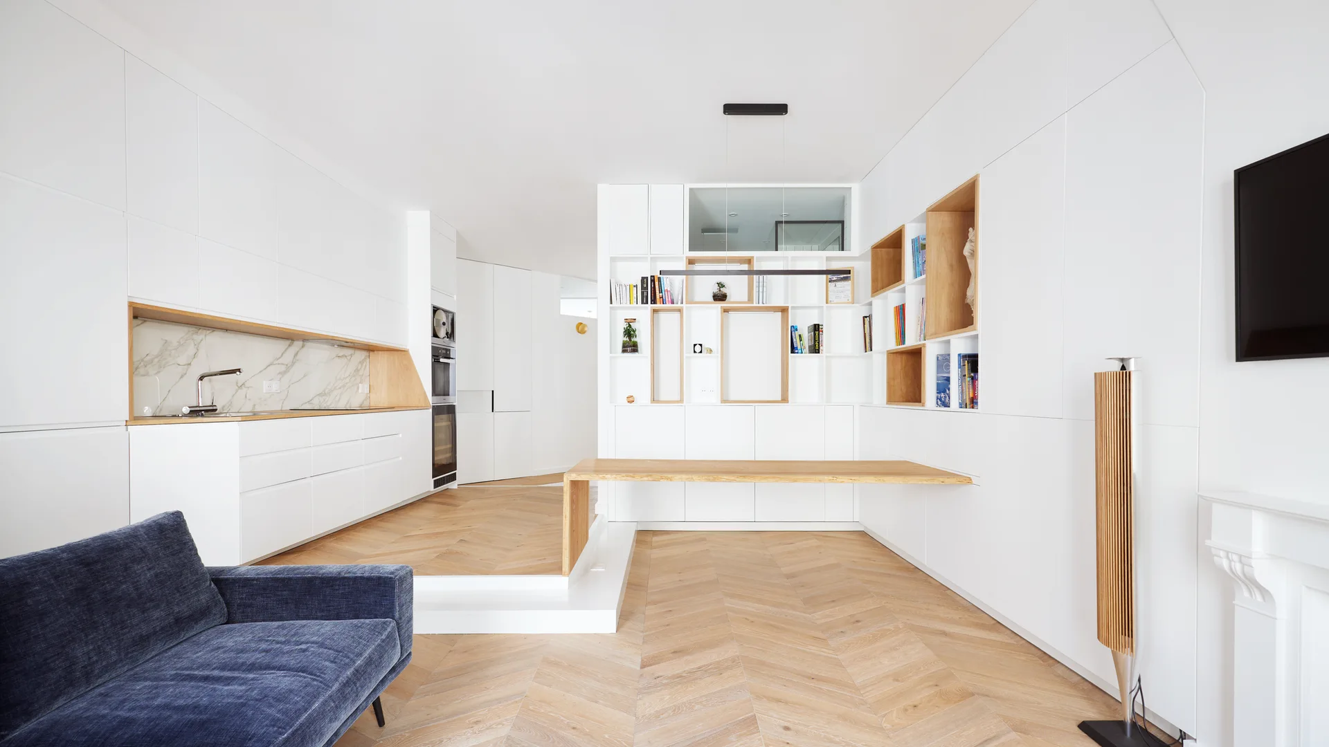Minimalist kitchen and living space featuring seamless white cabinetry, a light wood herringbone floor, and a floating wooden table.