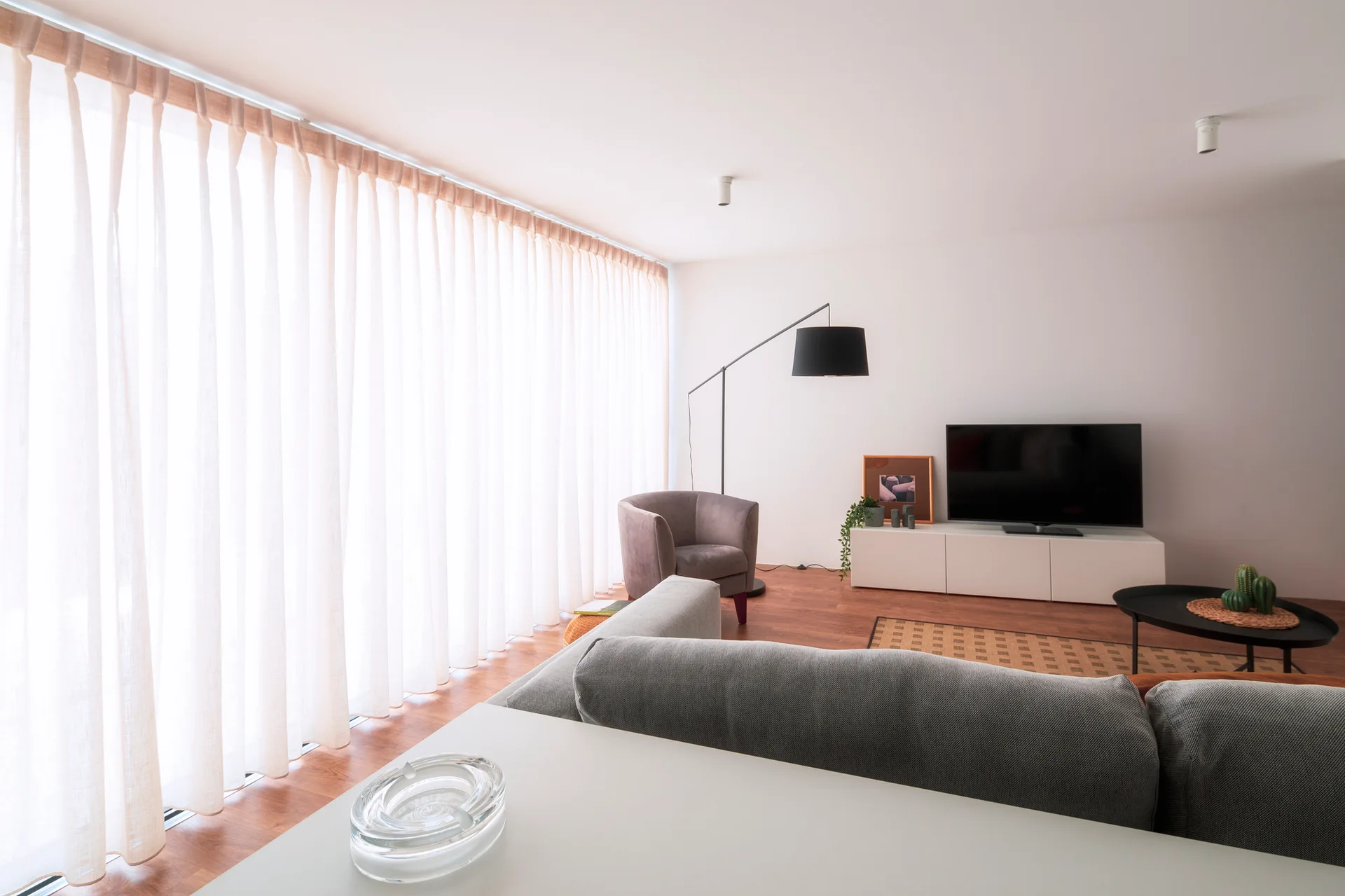 A bright living room with a gray sofa, low white TV console, and a large window wall covered by sheer floor-to-ceiling curtains.
