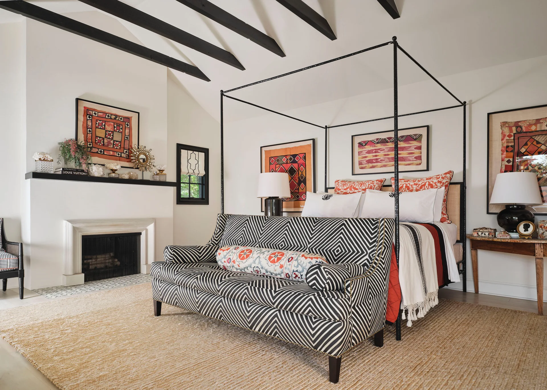 An eclectic bedroom featuring a black metal canopy bed, a black-and-white chevron sofa, dark ceiling beams, and framed textile art.
