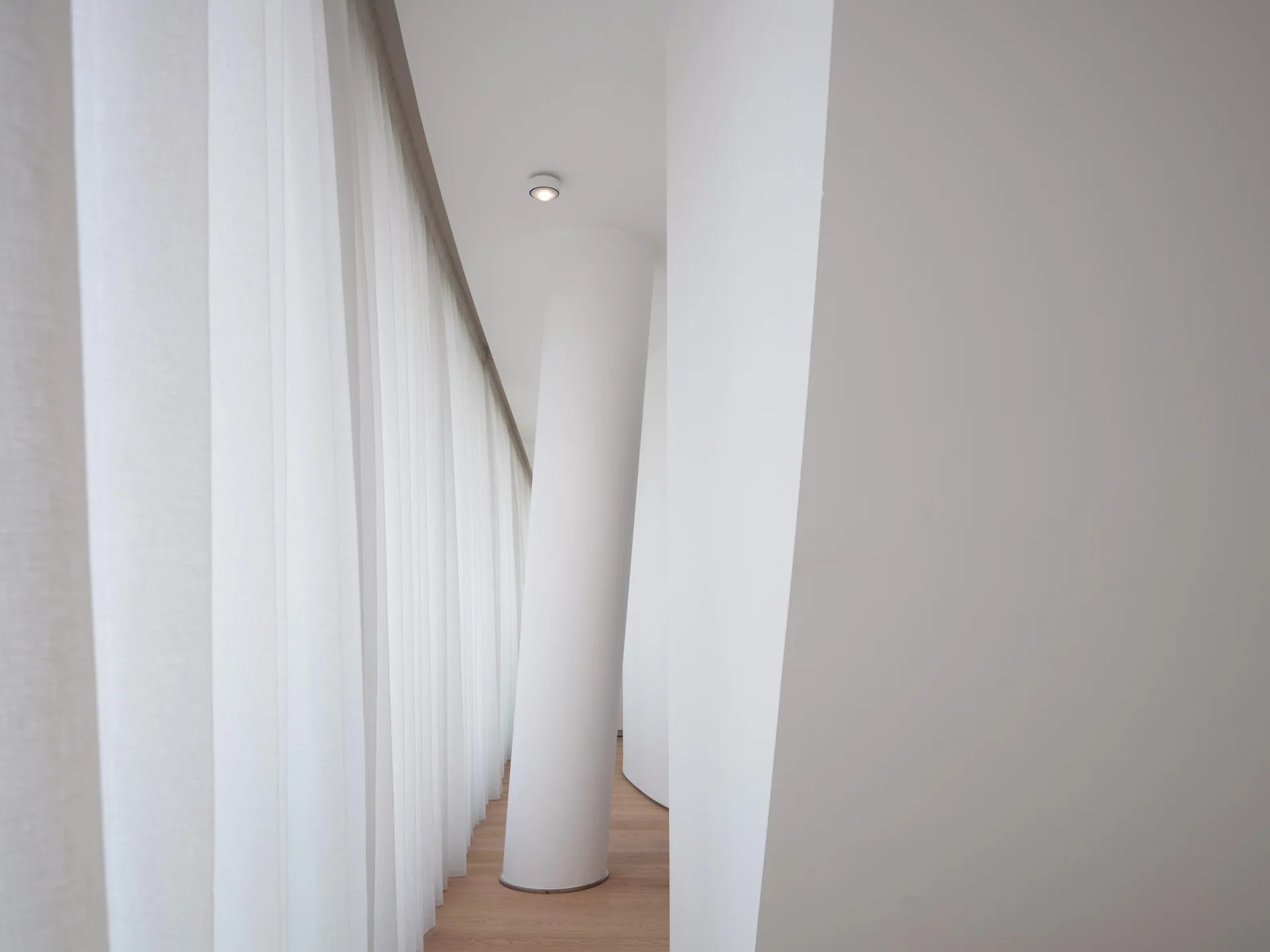 A minimalist white corridor featuring a thick cylindrical pillar, curved walls, light wood floors, and full-length white sheer curtains.