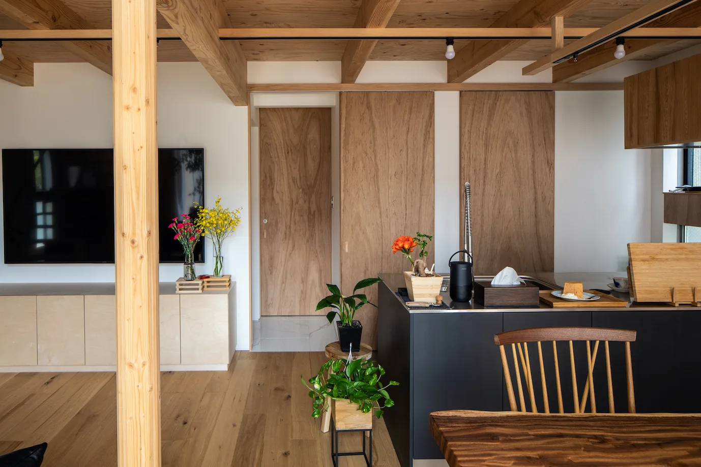 A minimalist residential kitchen and living space featuring exposed wood ceiling beams, vertical wood-paneled sliding doors, and a black island.