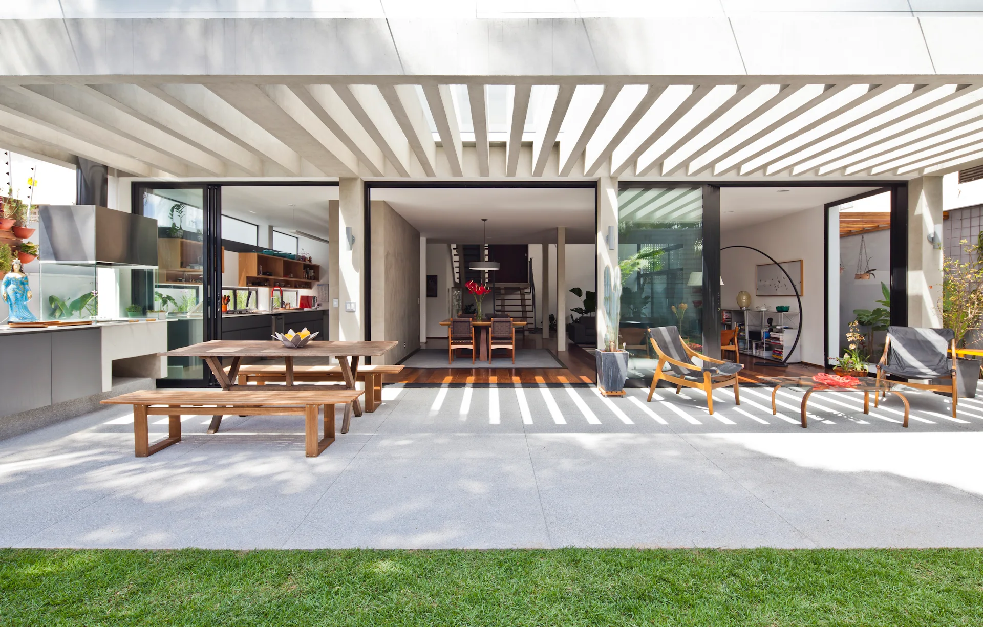 A modern gray concrete patio features a slatted overhead pergola, a wooden dining table with benches, and large sliding glass doors leading indoors.