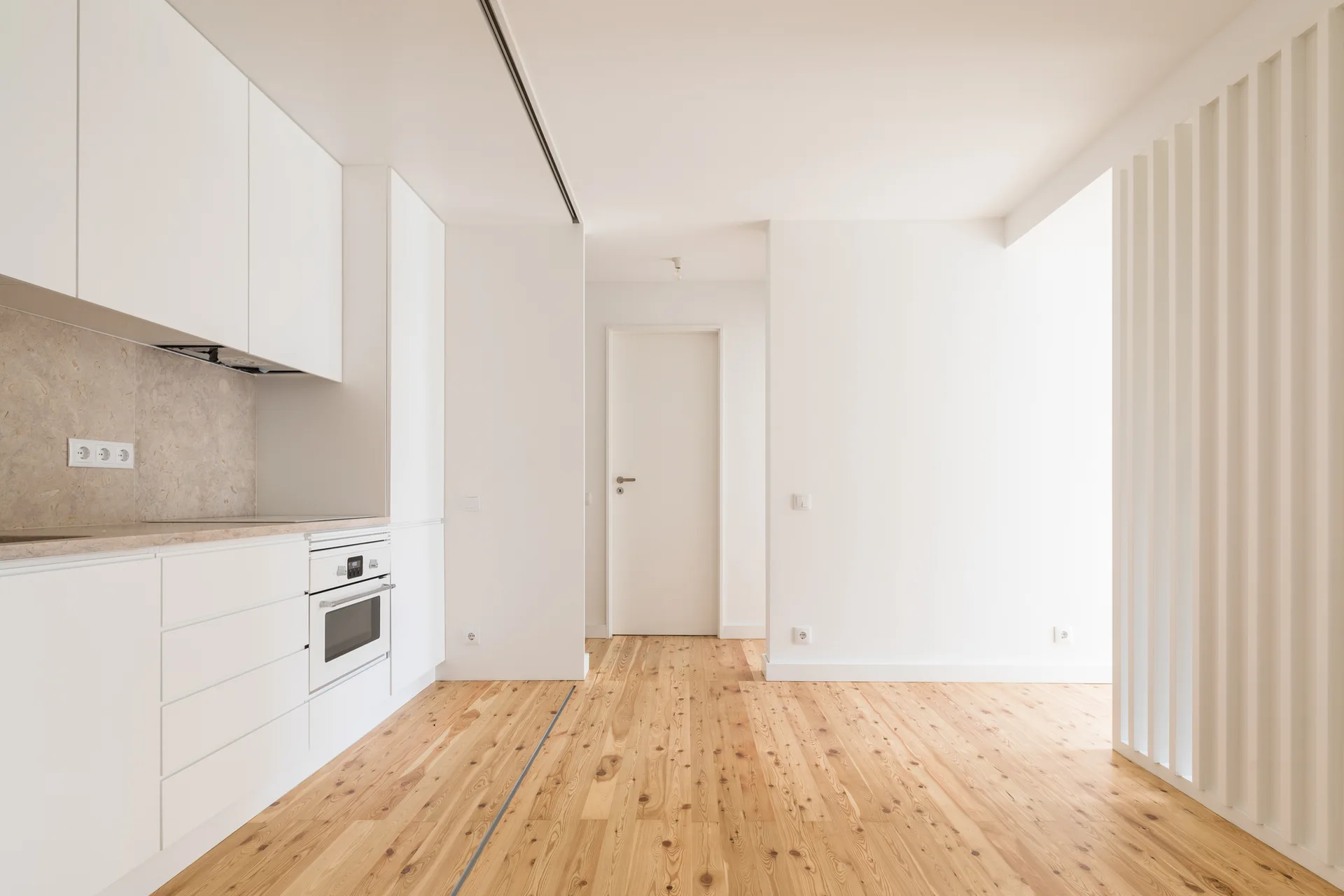 A minimalist white kitchen features flat-front cabinetry, a stone backsplash, and warm pine wood flooring alongside a white slatted partition wall.