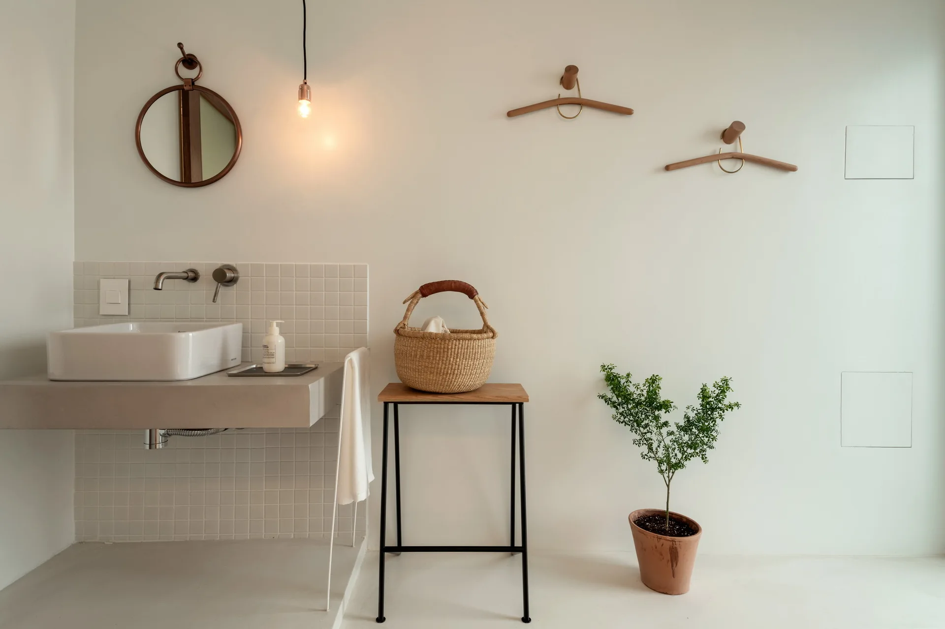 A minimalist bathroom features a wall-mounted concrete vanity with a white vessel sink, square white tiled backsplash, and wooden accents on white walls.