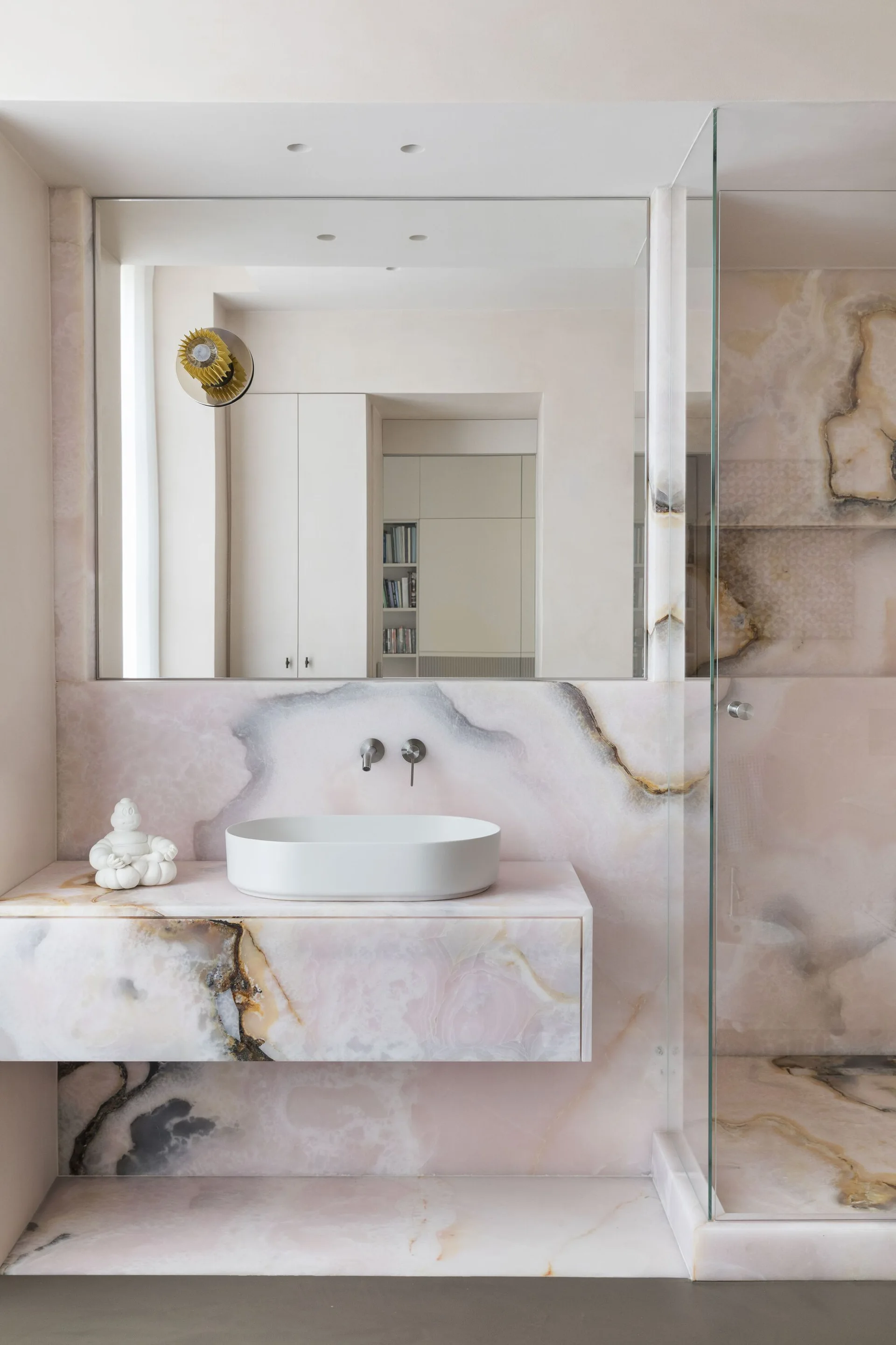 A minimalist bathroom features a floating pink onyx vanity with a white vessel sink, a large wall mirror, and matching veined stone cladding.