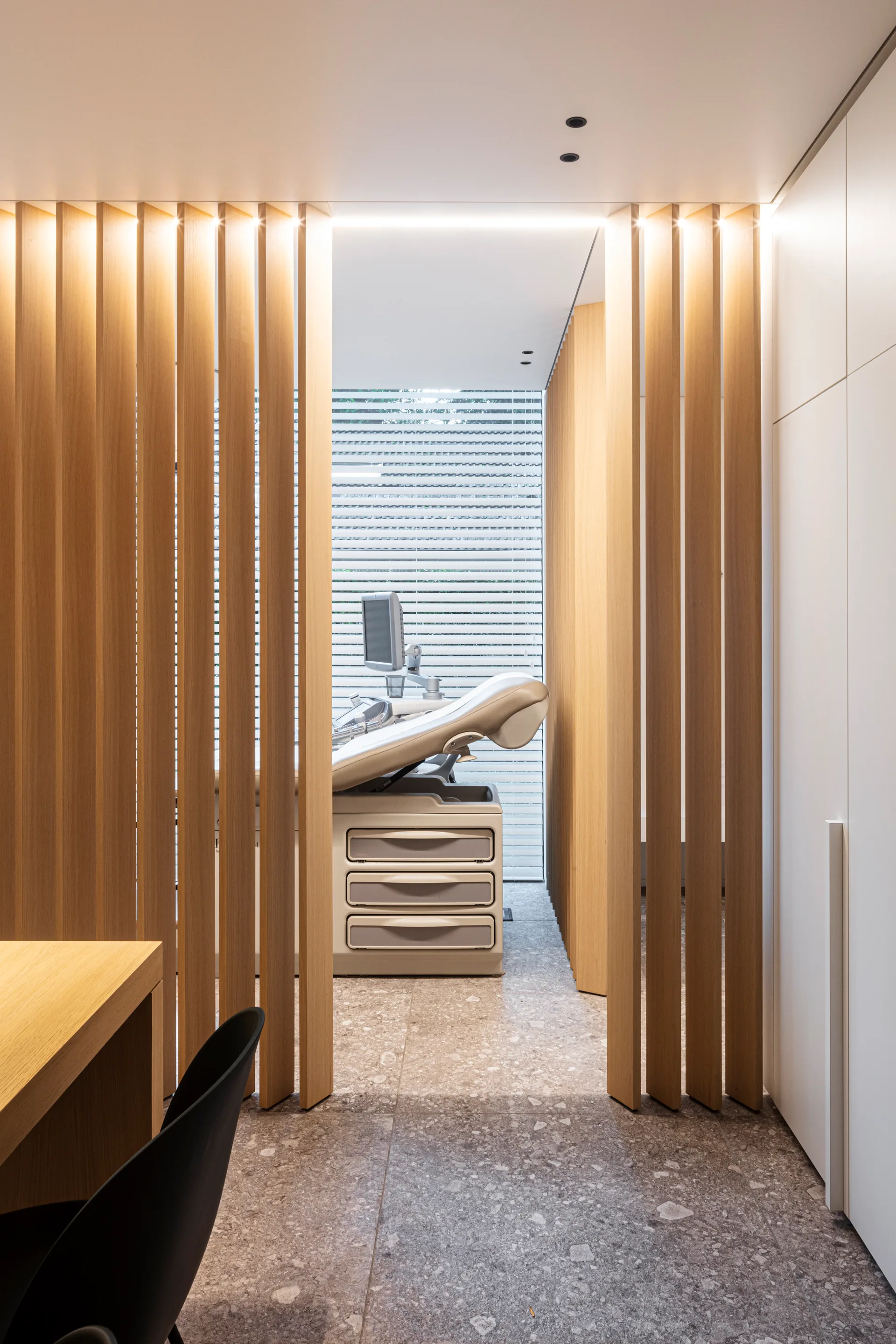 A vertical wood slat partition divides a desk area from a clinical exam space with speckled stone flooring and seamless white cabinetry.