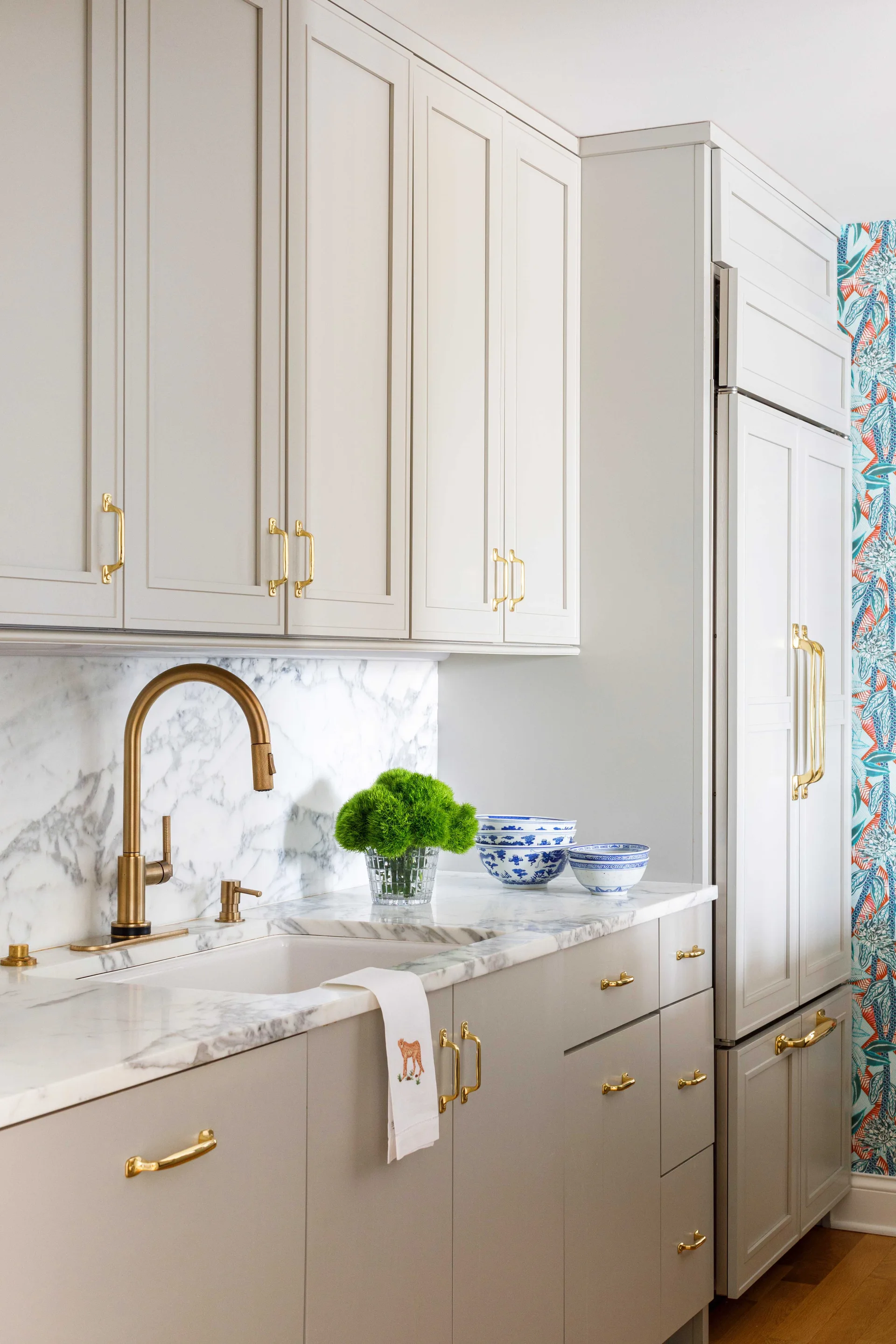 A kitchen features light beige shaker cabinetry with brass handles, a veined white marble backsplash, and an integrated refrigerator.
