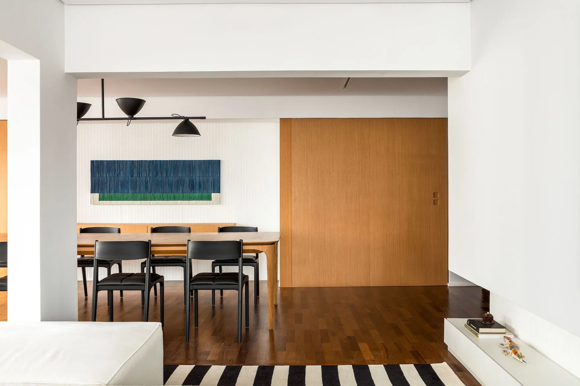 A minimalist dining room features a wooden dining table with black chairs, a fluted white accent wall, and a large wood sliding door.