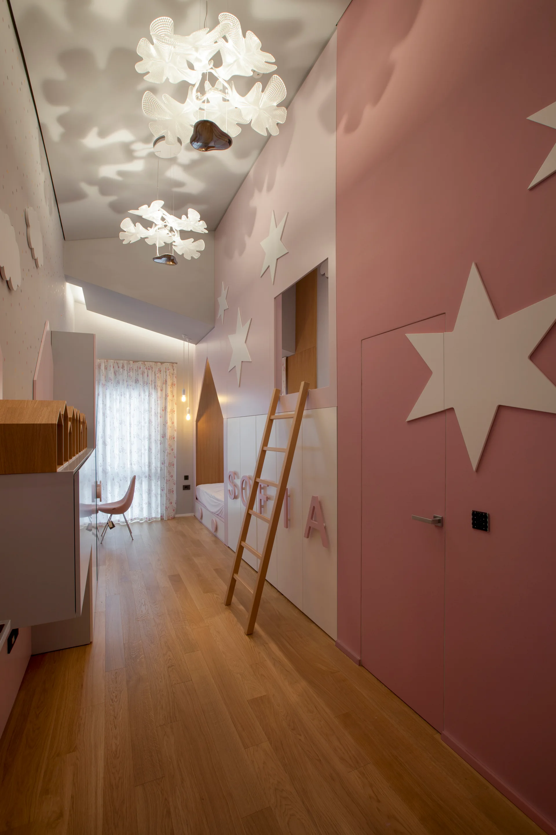 A children's bedroom features pink paneled walls decorated with white stars, custom storage with a wooden ladder, and sculptural white ceiling lights.