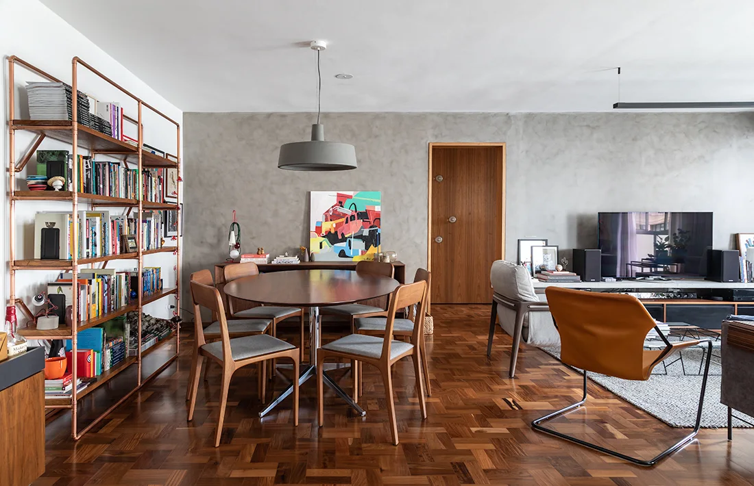 An open living and dining area with parquet floors, a grey concrete feature wall, a copper-pipe bookshelf, and a round wooden dining set.